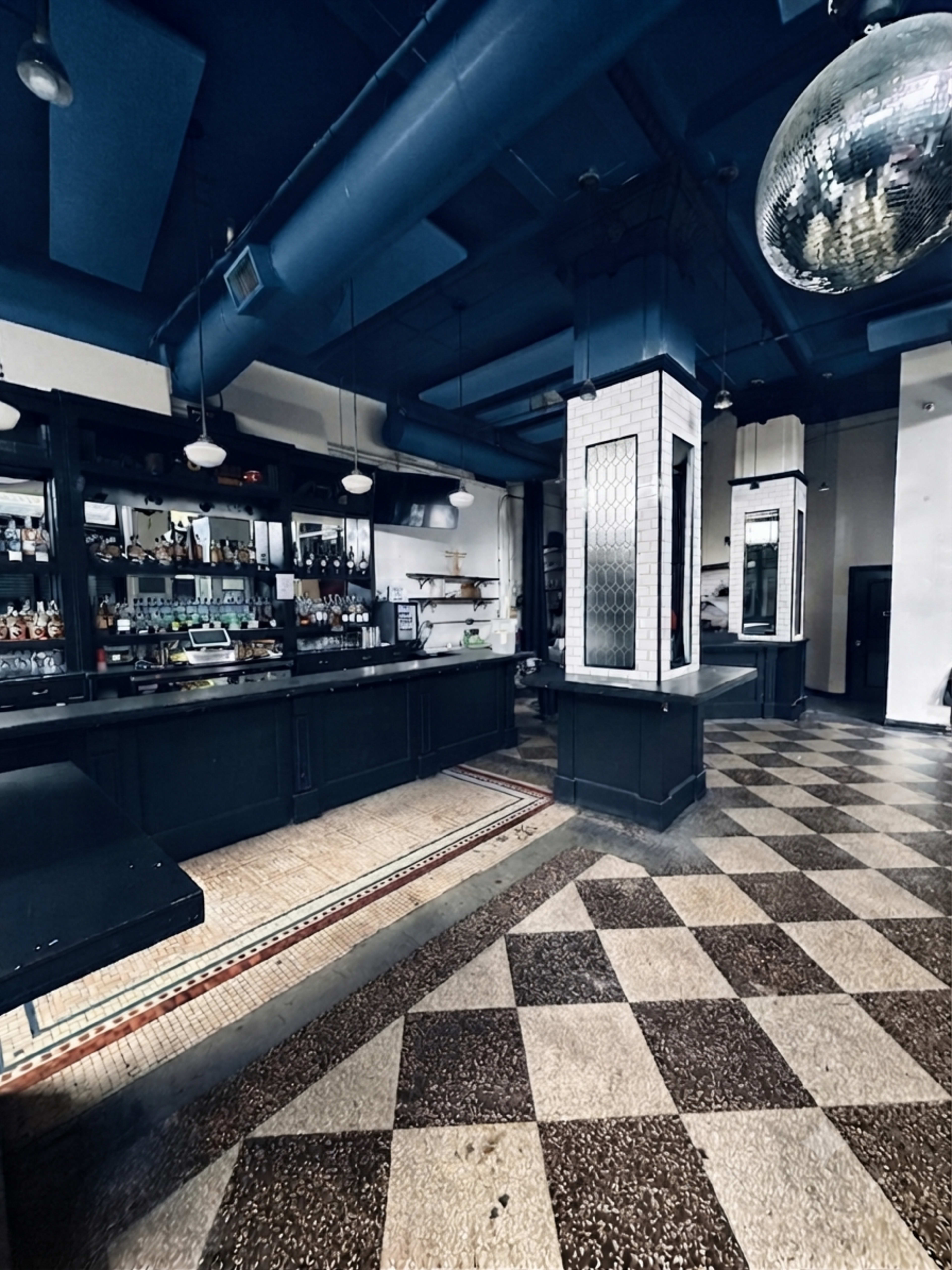 The interior of a bar features dark wood counters, a mirrored wall, and a checkered tile floor, with a large disco ball hanging from the ceiling.