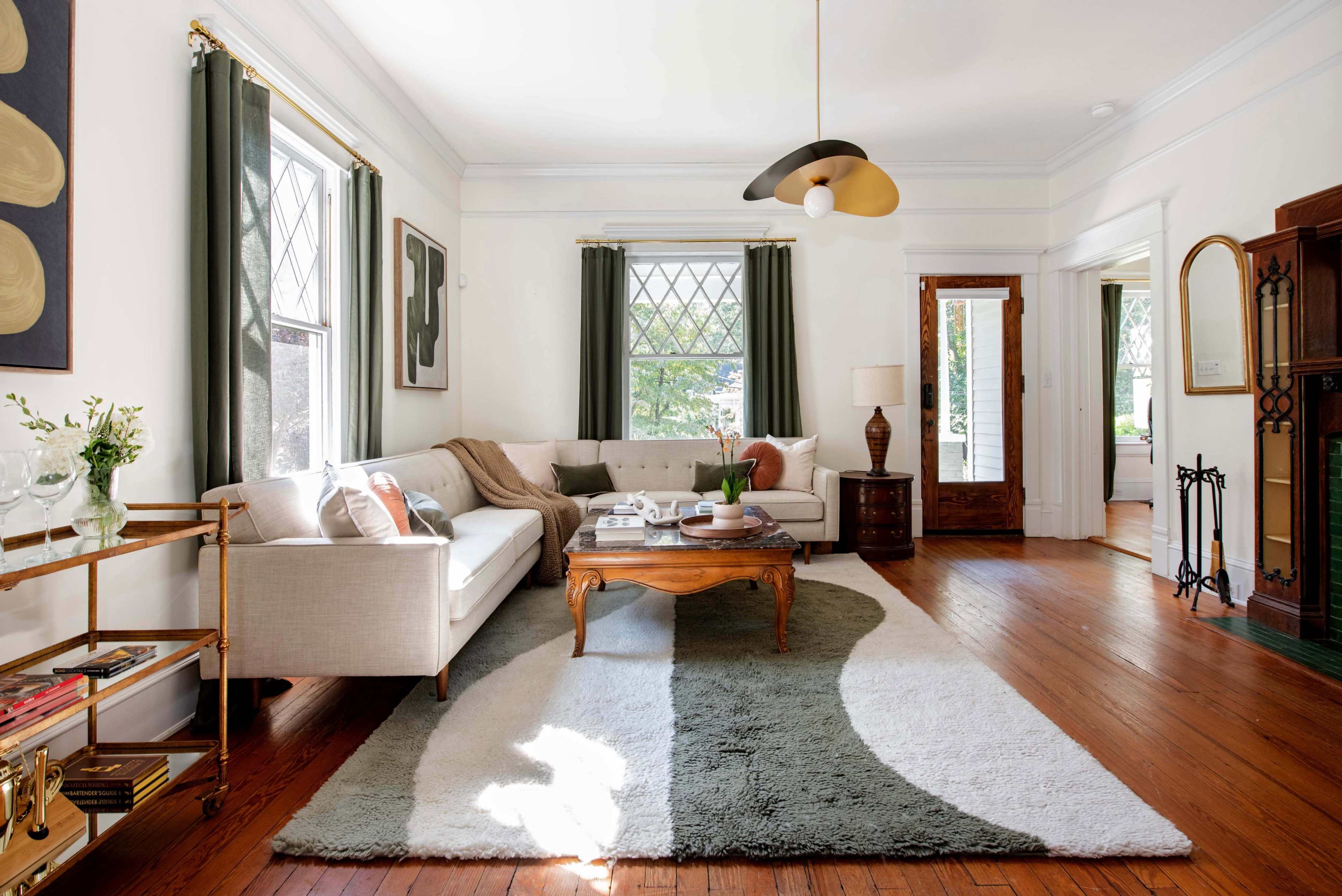 A bright living room features a beige sofa, a round coffee table, and a large area rug, complemented by green curtains and hardwood floors.