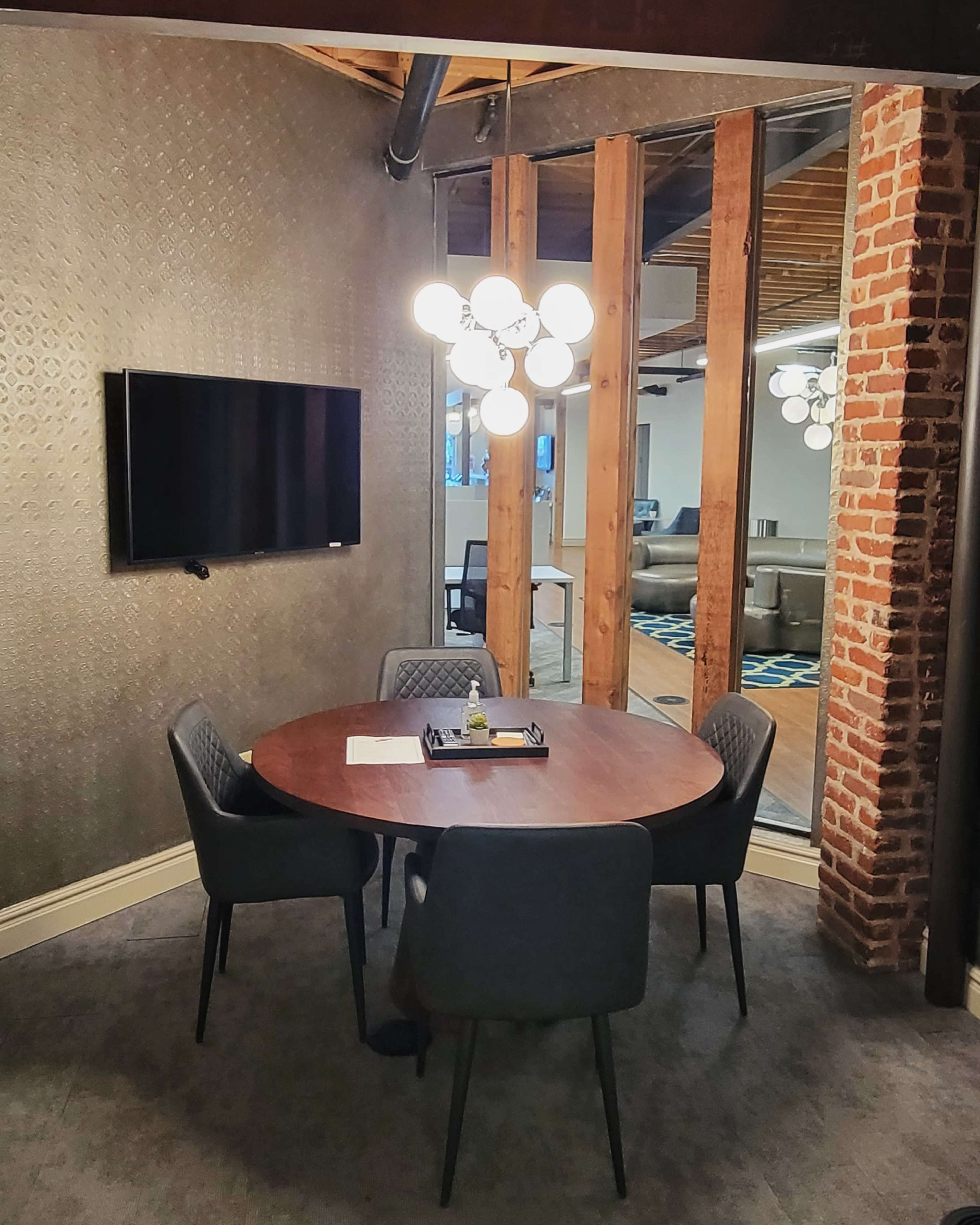 A small conference room features a round wooden table with four chairs, a television mounted on the wall, and a decorative light fixture.
