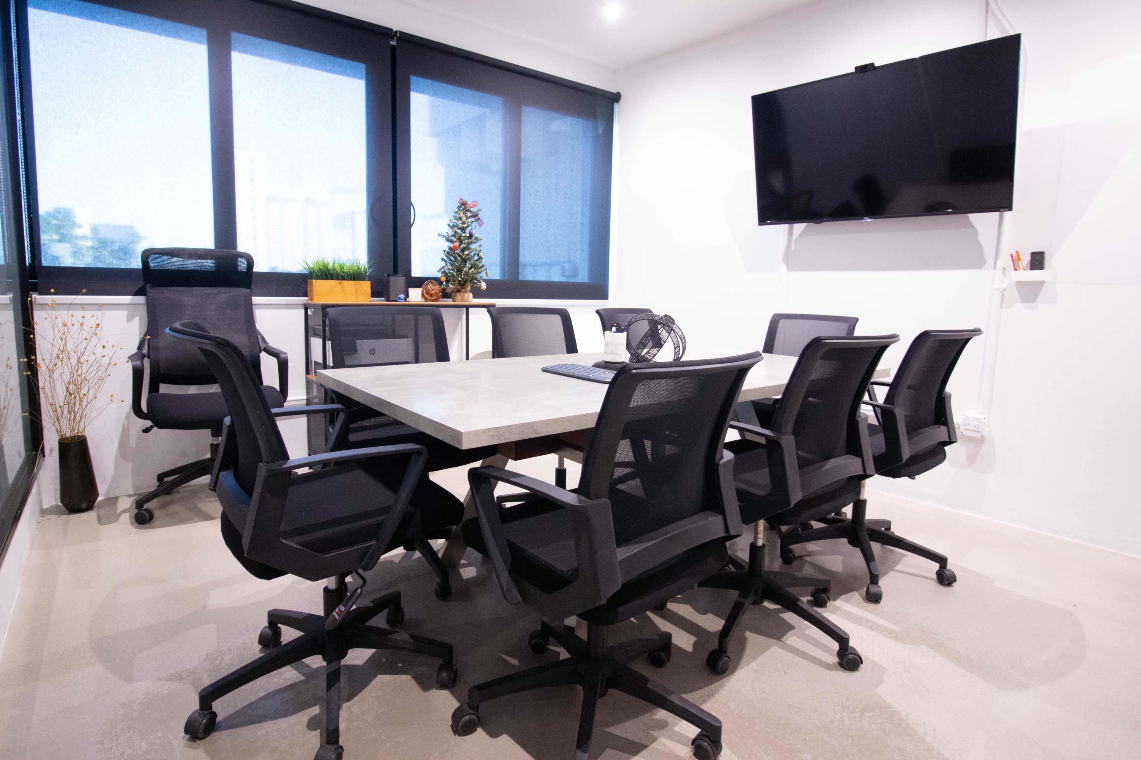 A modern conference room with a large table surrounded by eight black office chairs, a television mounted on the wall, and windows letting in natural light.