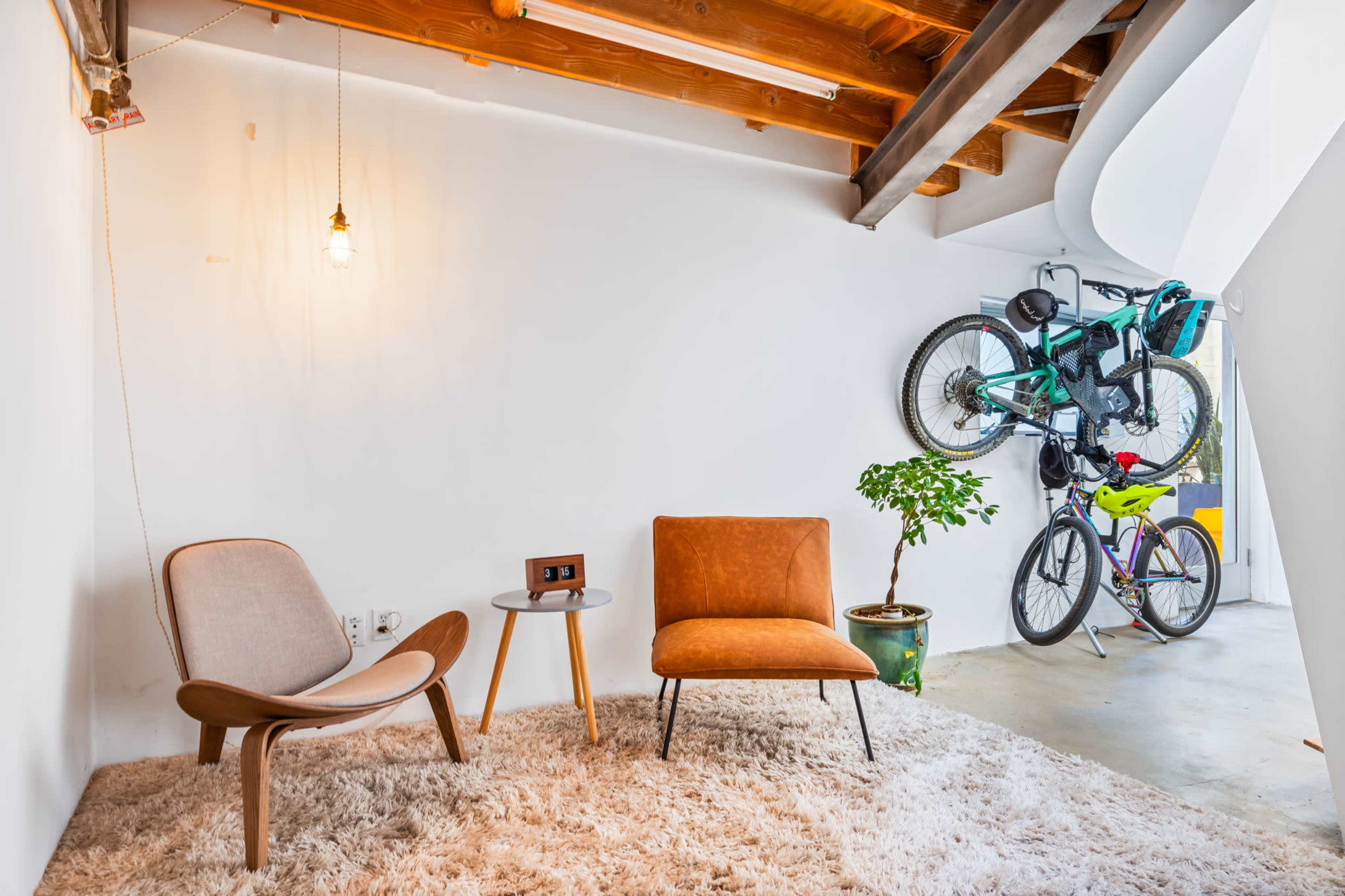 A modern living space featuring two chairs on a plush rug, a side table with a clock, a potted plant, and two bicycles mounted on the wall.