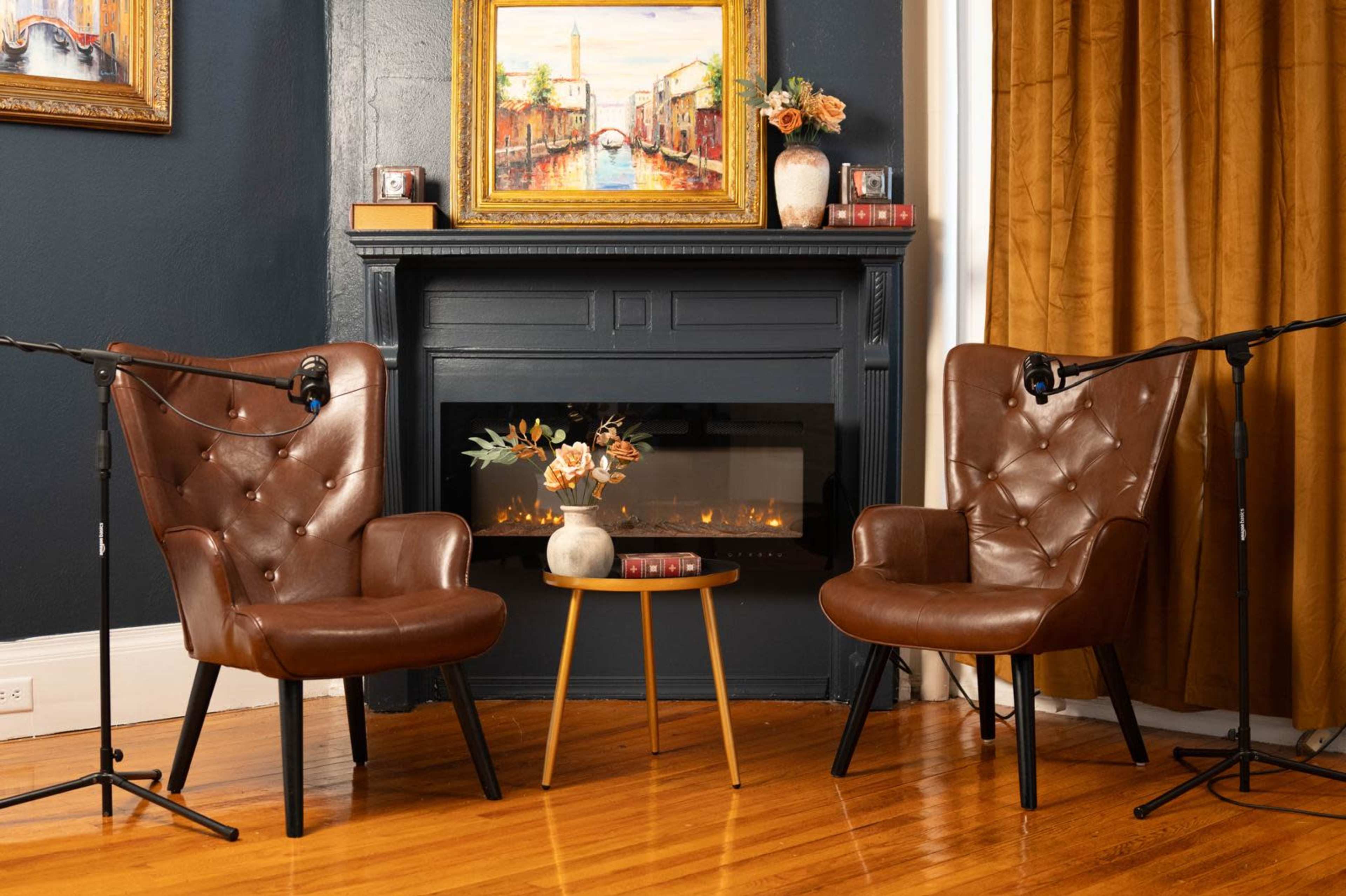 Two brown leather armchairs positioned around a small round table in front of a fireplace, with a painting on the wall and microphones set up near the chairs.