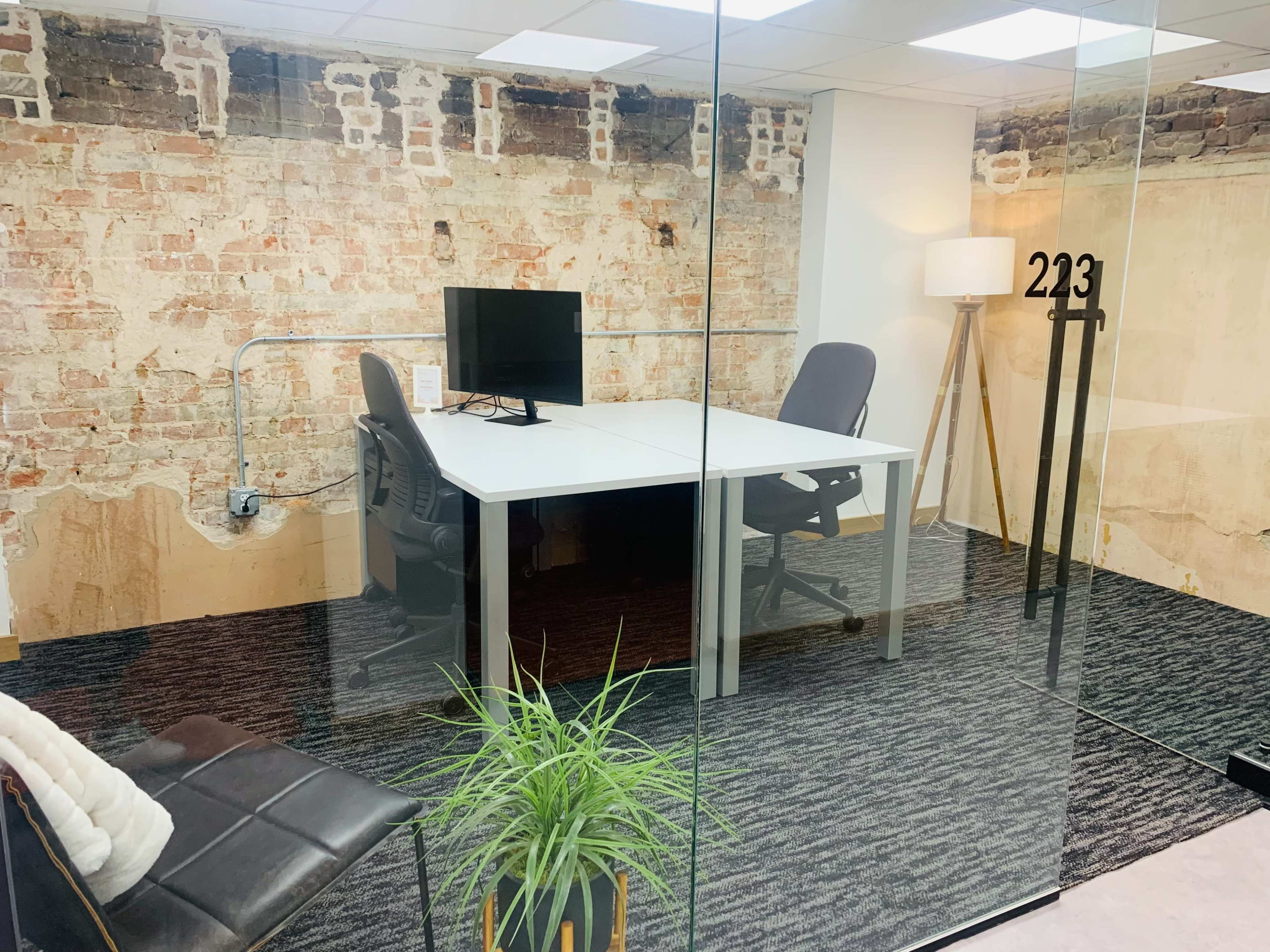 A modern office space with a glass wall, featuring a desk, a computer monitor, and a minimalist armchair, against a backdrop of exposed brick.