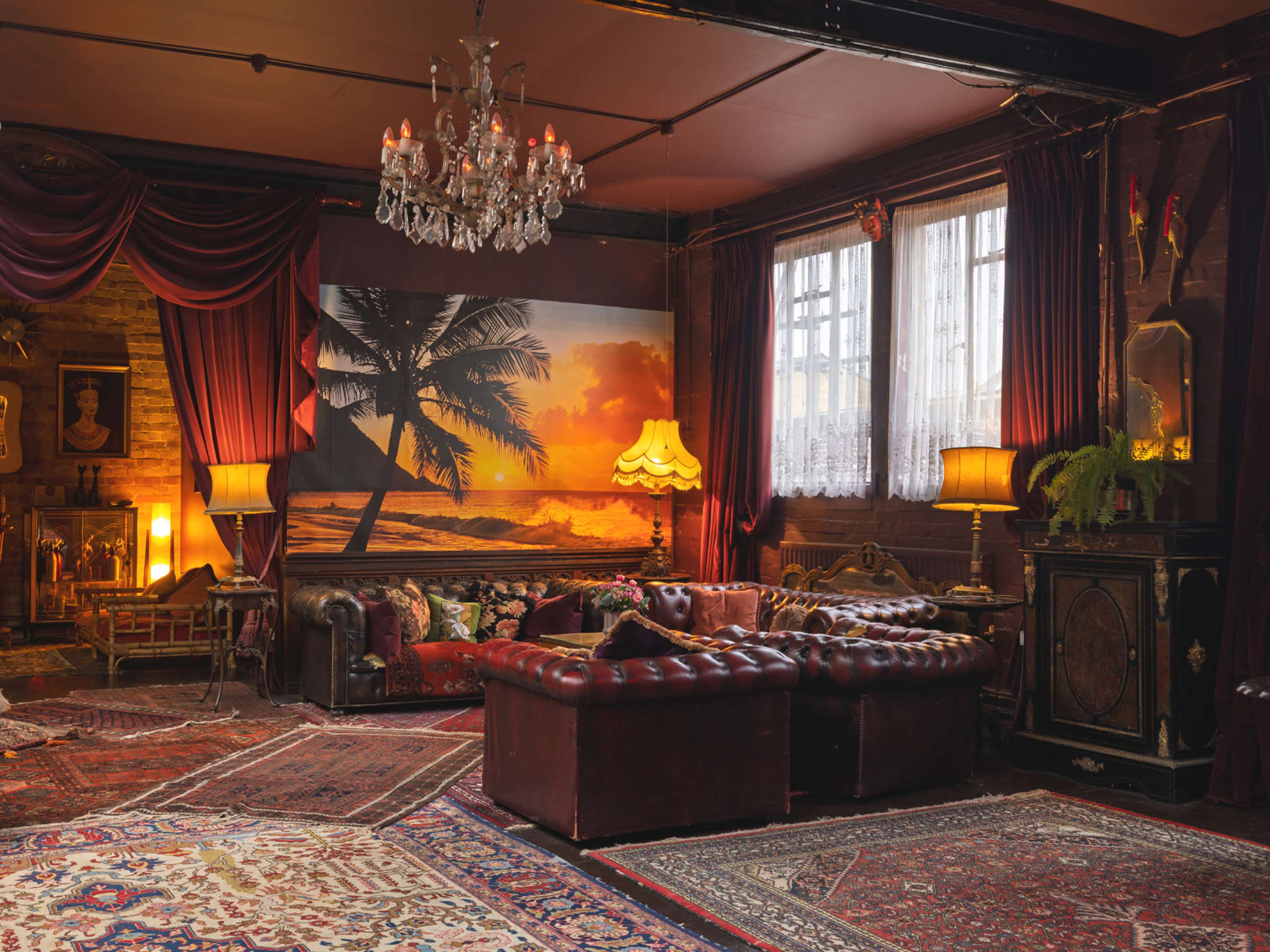 The room features a vintage leather sofa, ornate rugs, and a large mural of a sunset with palm trees, illuminated by table lamps and a chandelier.