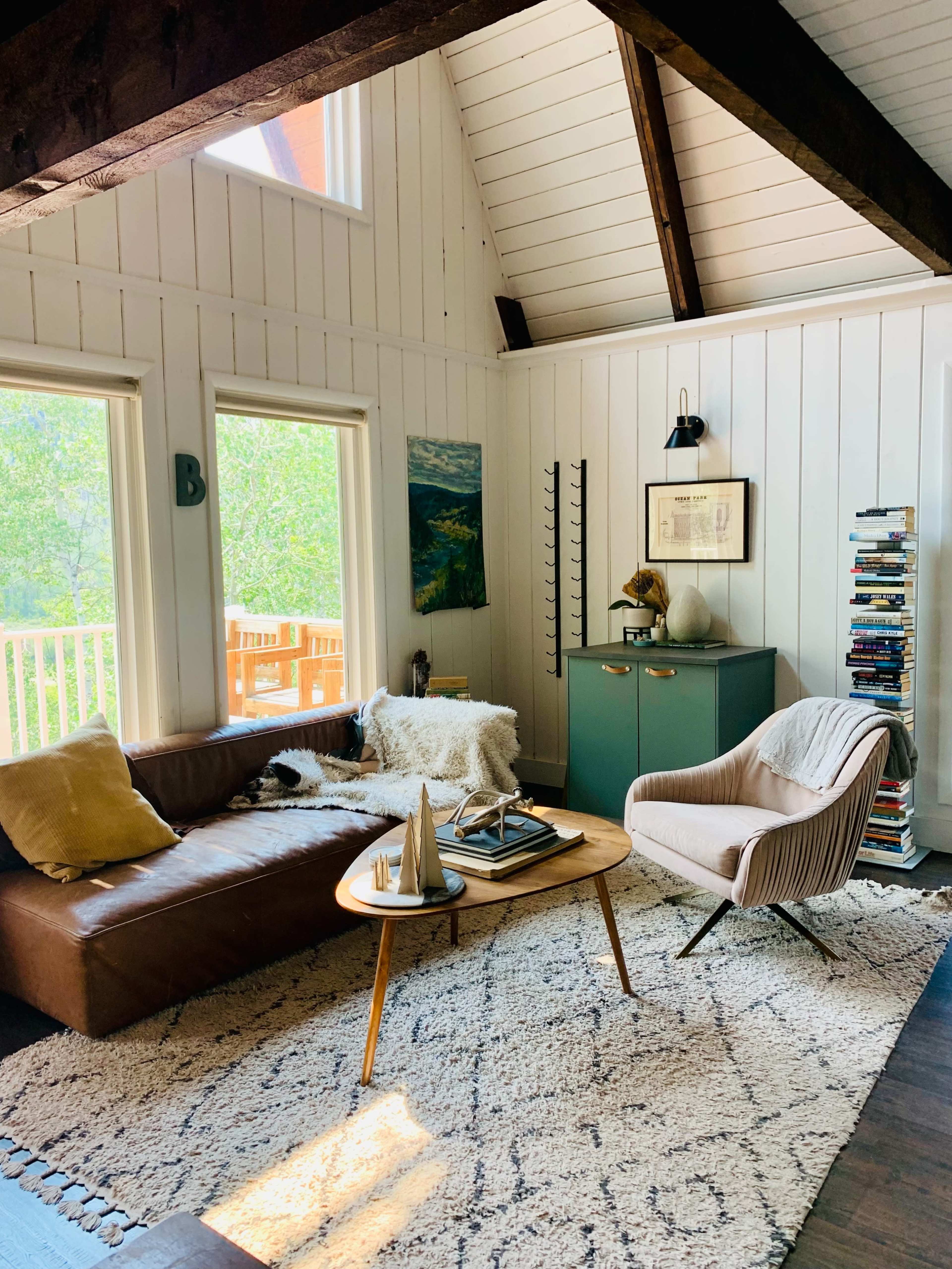 A cozy living room features a brown leather sofa, a round wooden coffee table, a pale-colored armchair, and a bookshelf, all set against a backdrop of large windows and exposed wooden beams.