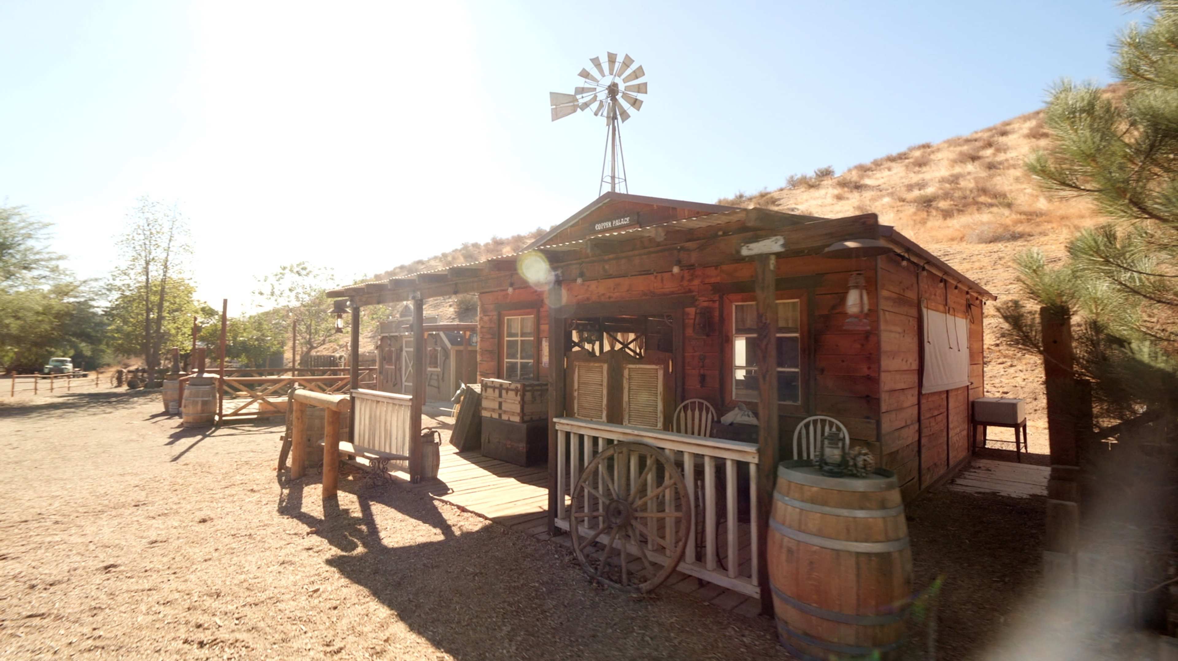 A rustic wooden building with a covered porch and a windmill stands in a dry landscape.