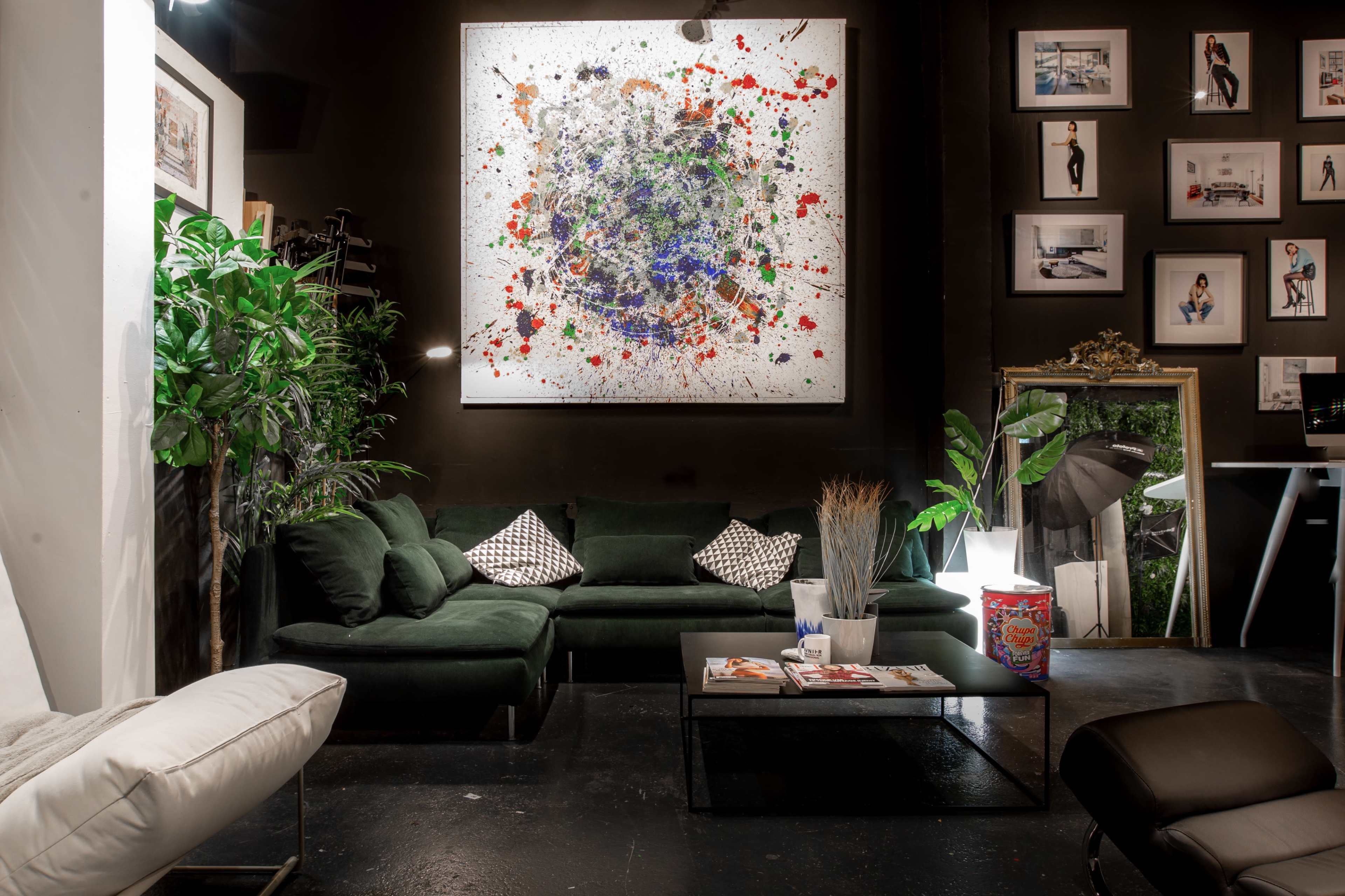 A modern interior with a green couch, a coffee table with magazines, and a large abstract painting on the wall, surrounded by various framed photographs and plants.