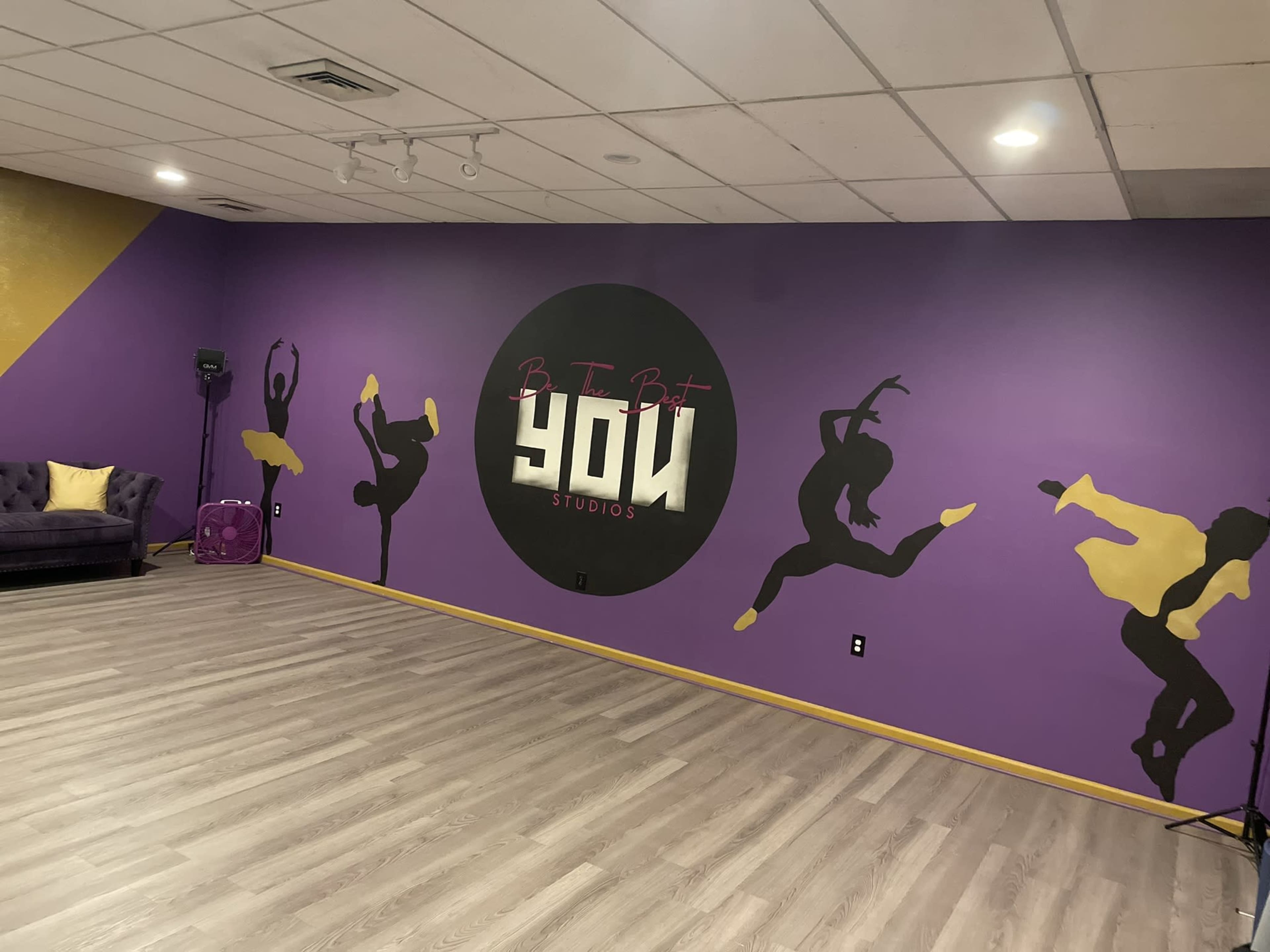 The image shows a dance studio with a purple and yellow wall featuring silhouettes of dancers and the words "Be It But YOU Studios."