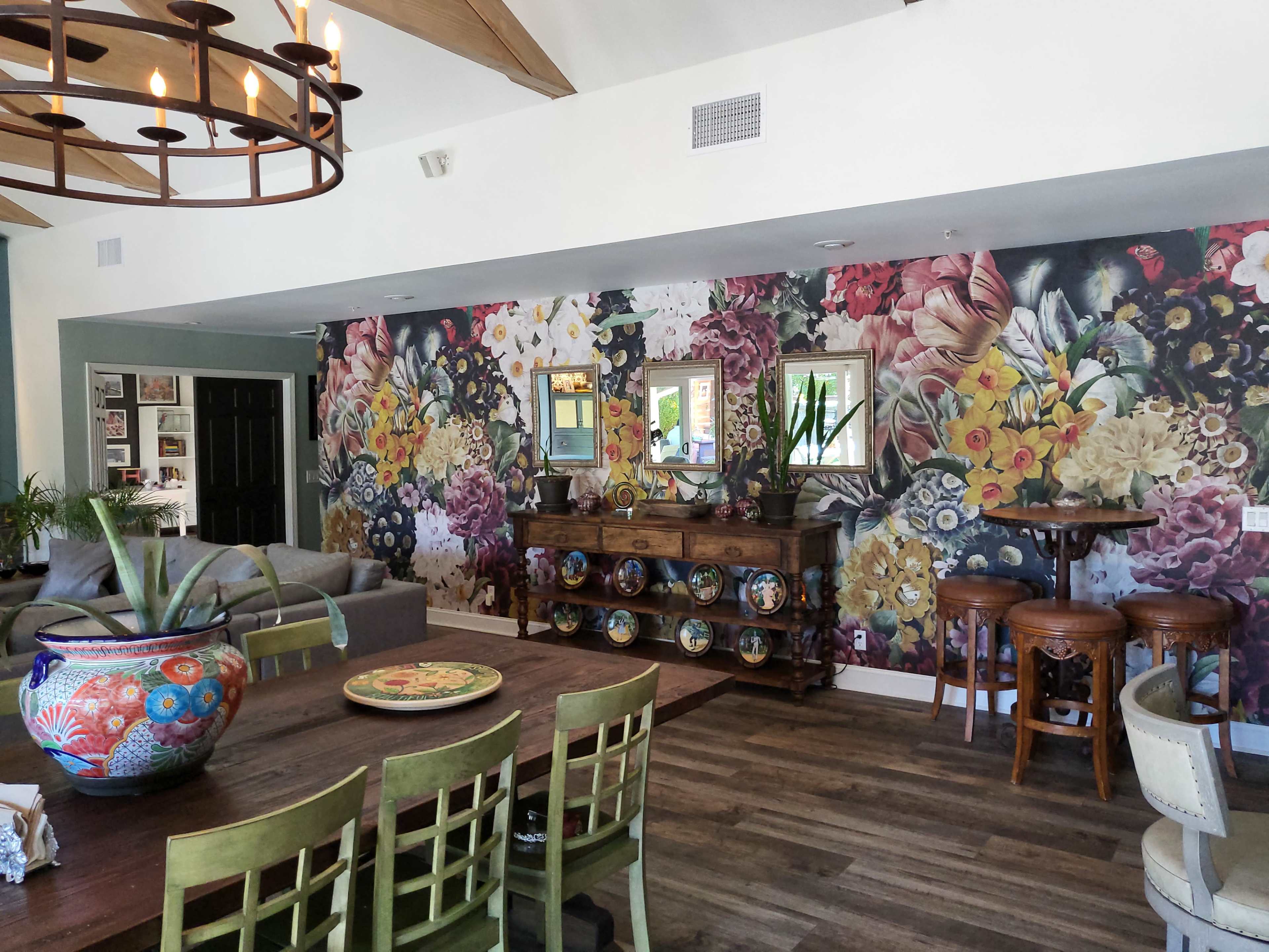 A brightly lit living space featuring a floral wallpaper accent wall, a wooden dining table with green chairs, and decorative pottery displayed on a sideboard.