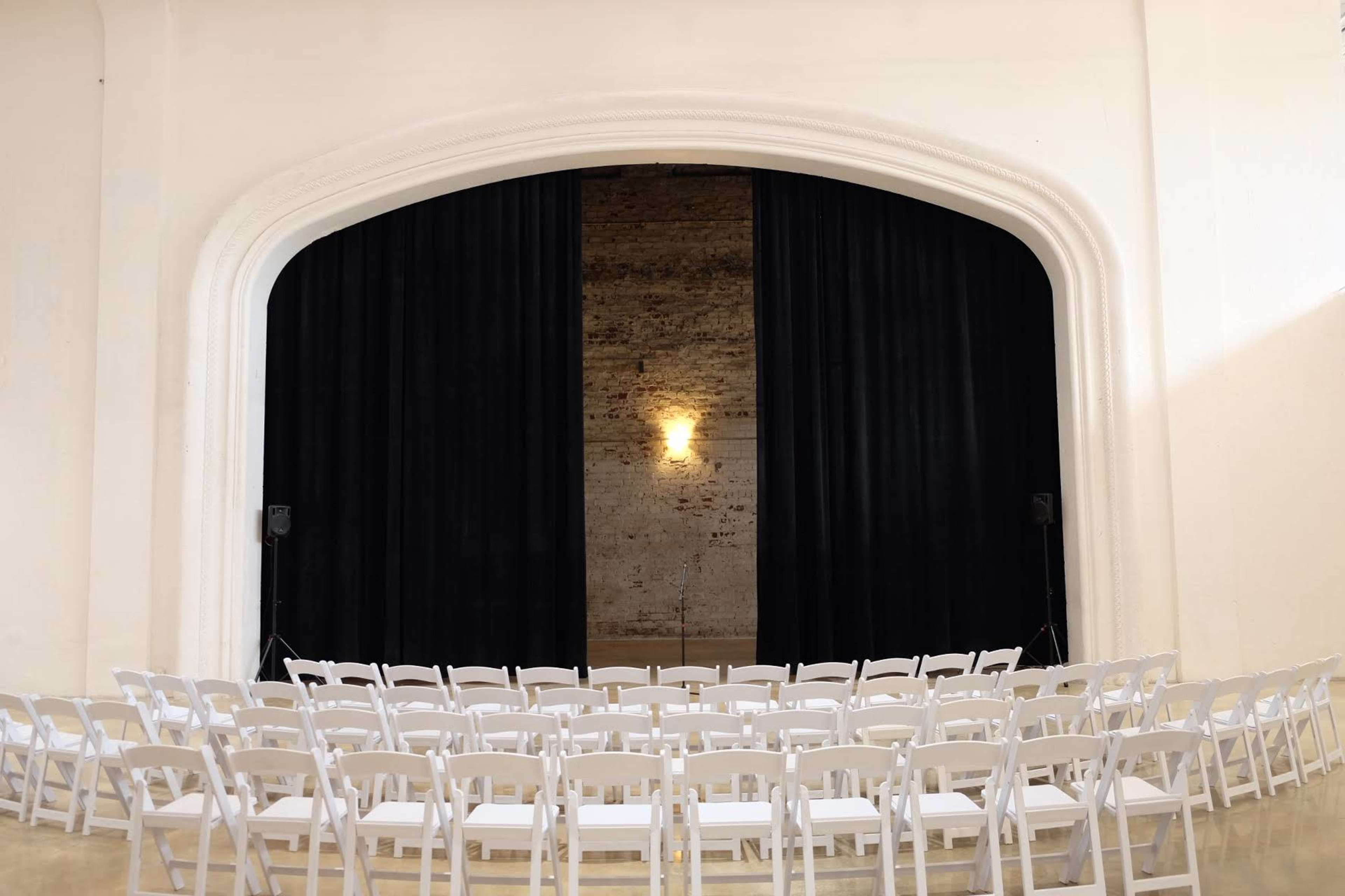 A row of white chairs is arranged in front of a stage with black curtains and a spotlight in a large, empty room.