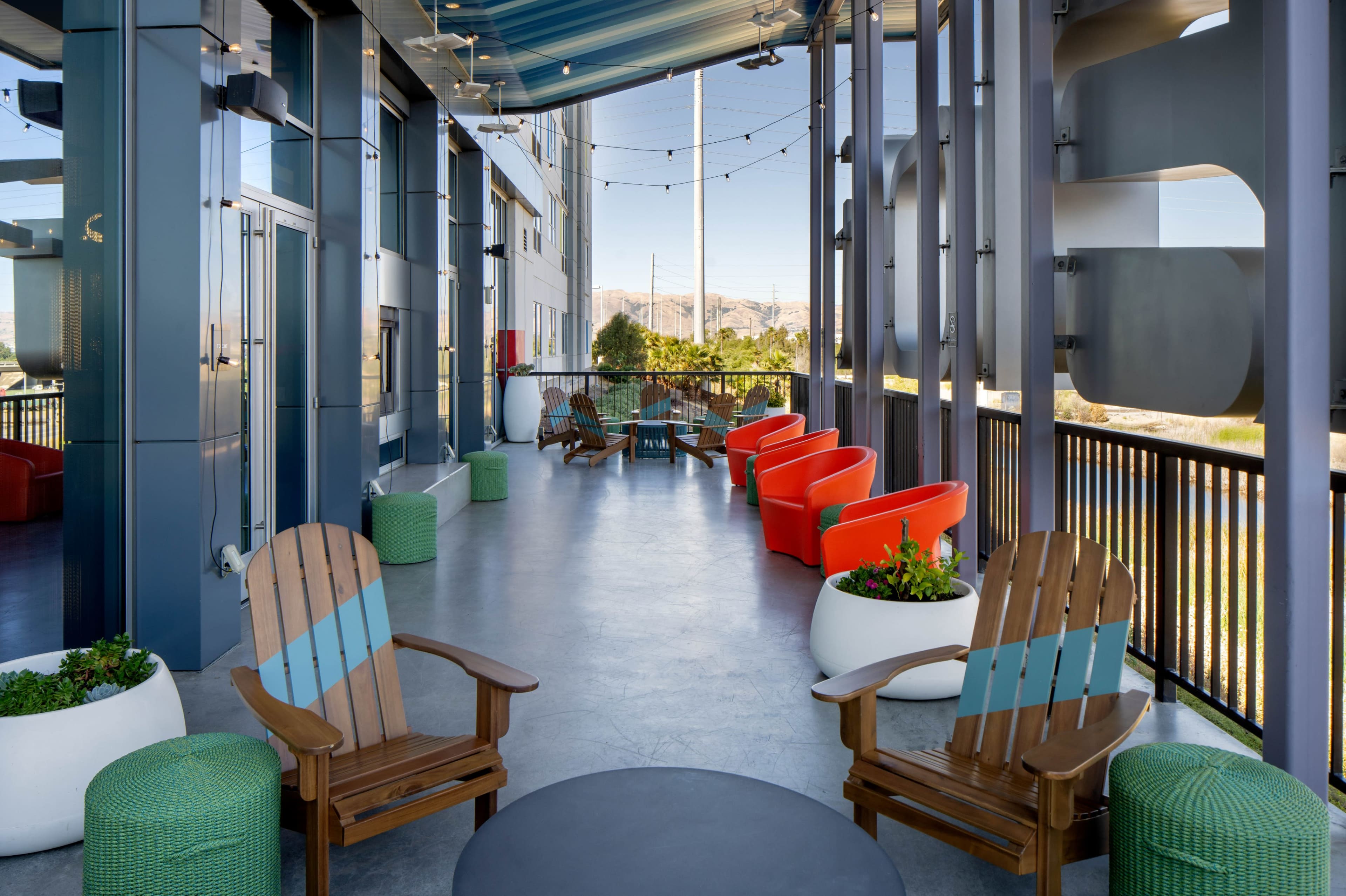 A modern outdoor lounge area with wooden chairs, vibrant orange seating, and planters, overlooking a landscape with mountains in the background.