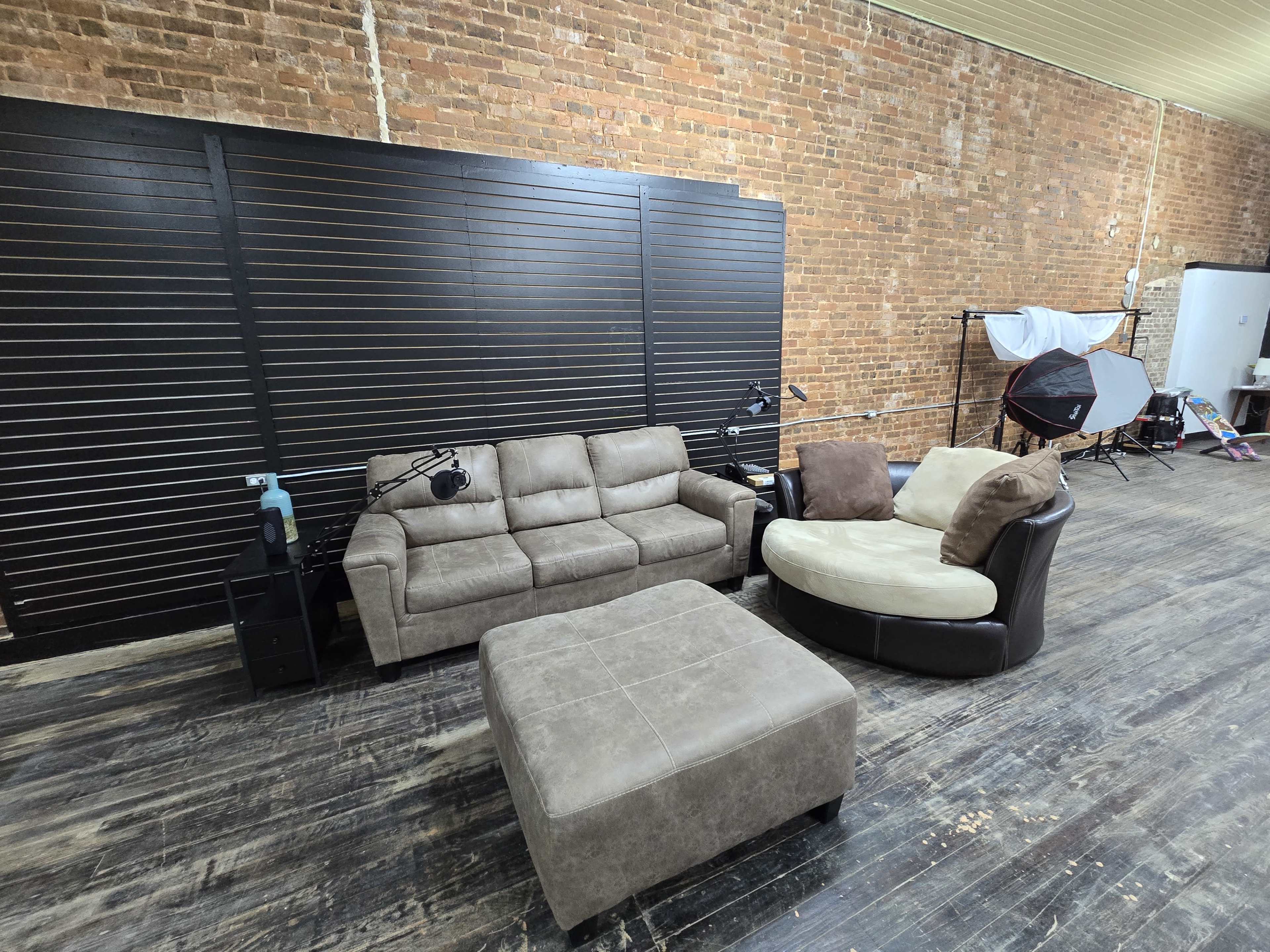 The image shows a seating area with a brown sectional sofa, a circular armchair, and a square ottoman on a wooden floor in a space featuring exposed brick walls.