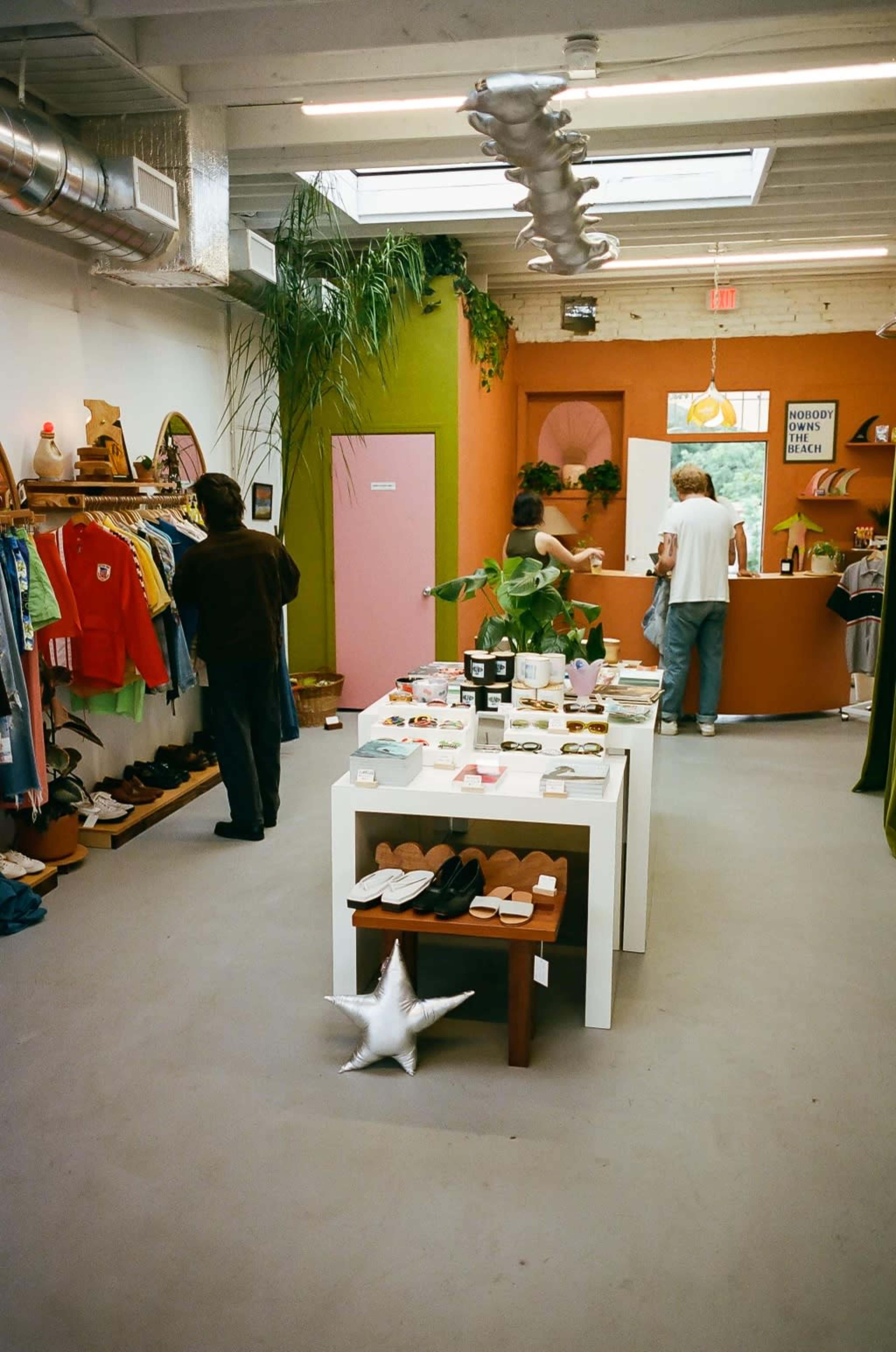 A retail store displays clothing and accessories on shelves and tables, with customers browsing in the background.