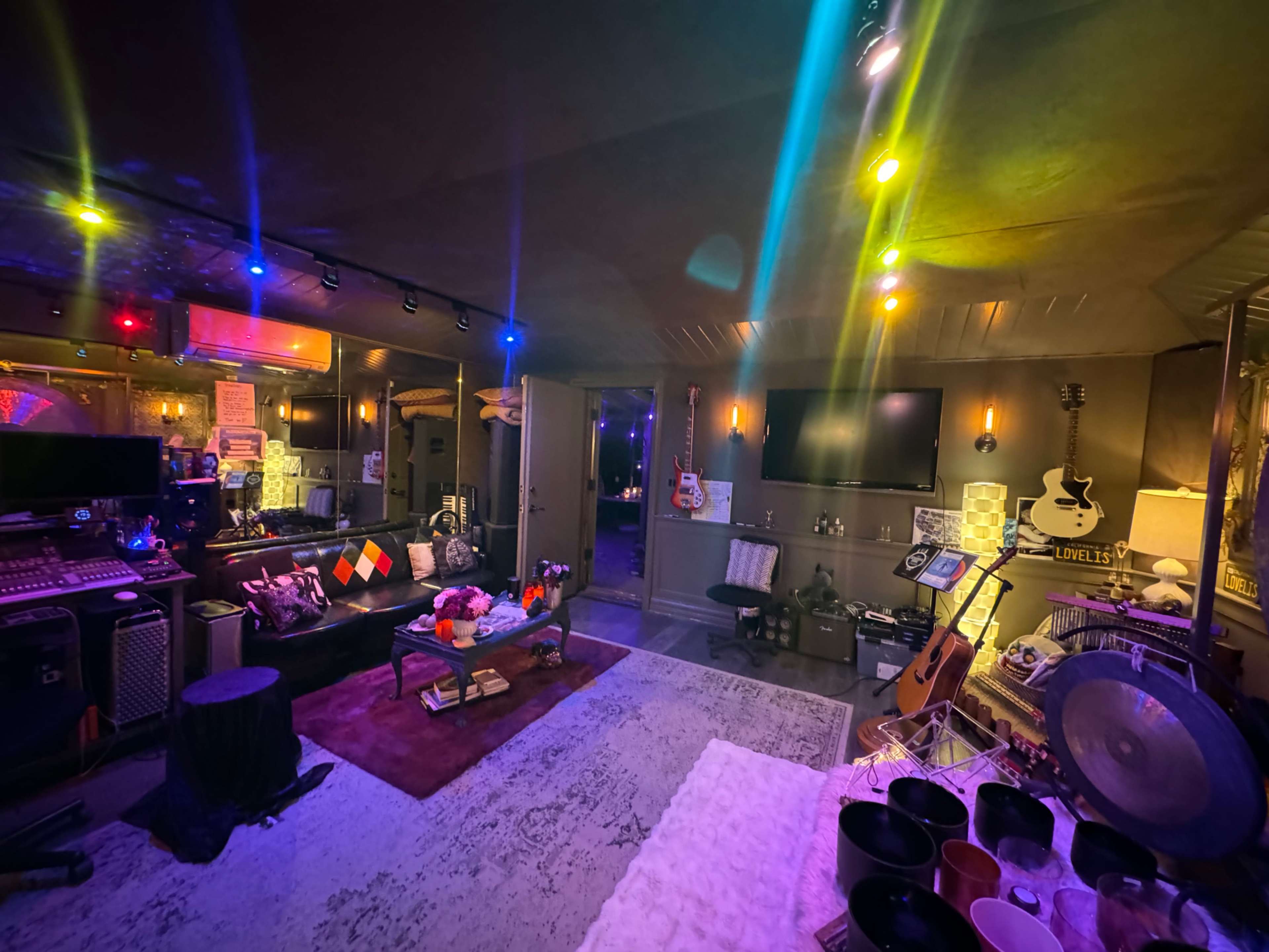 The image shows a music studio or rehearsal space with colorful lighting, various musical instruments, and a cozy seating area.