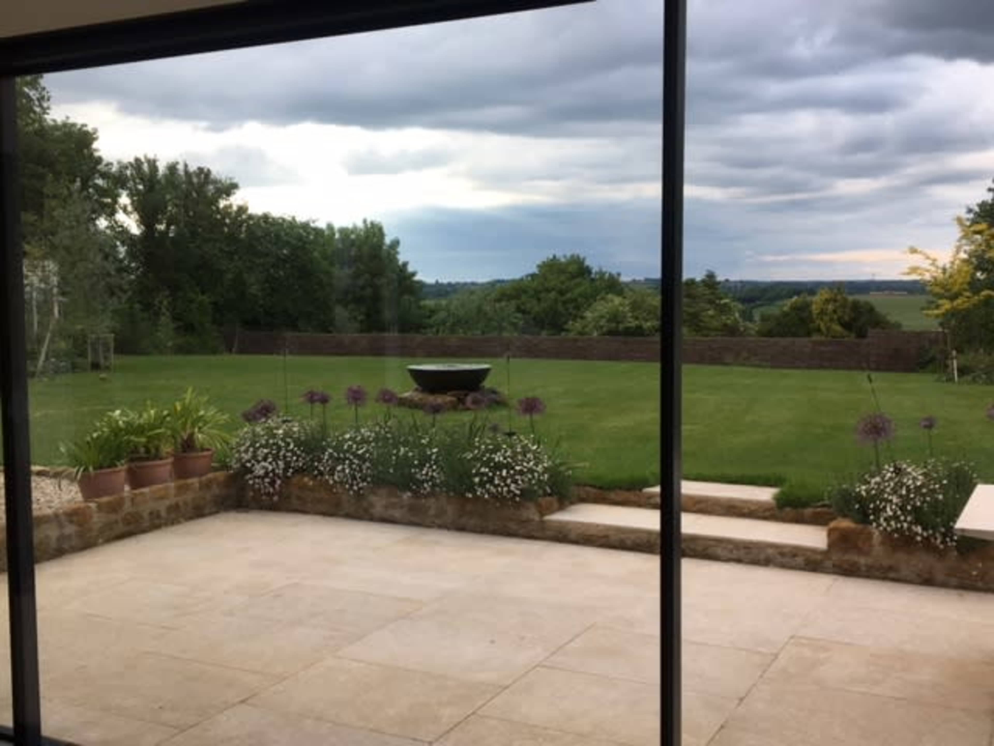 A patio with stone steps leads to a grassy yard surrounded by trees and flowers, under a cloudy sky.