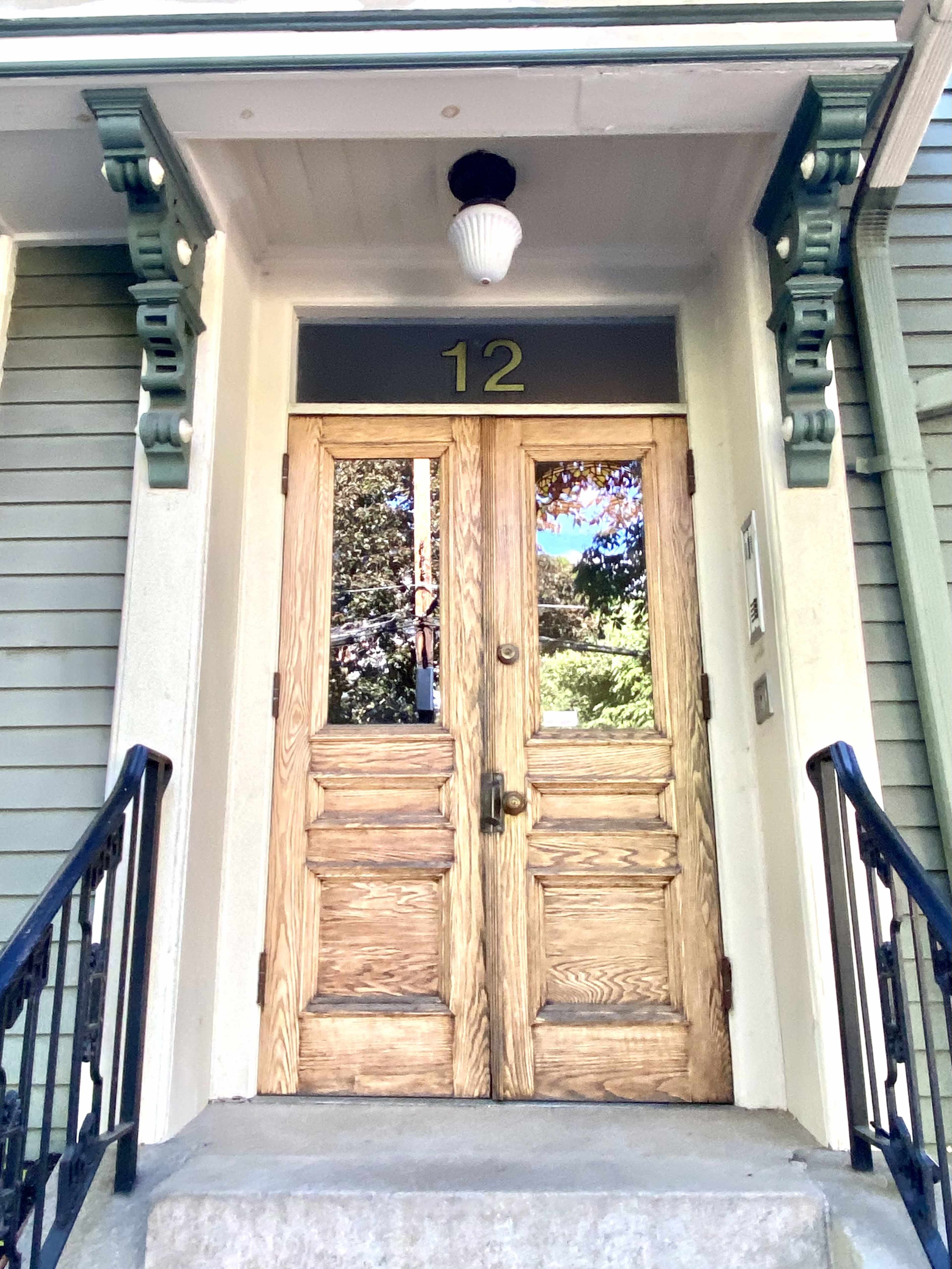 A pair of wooden double doors with a brass doorknob and the number "12" displayed above, set within a building featuring a green wooden exterior.