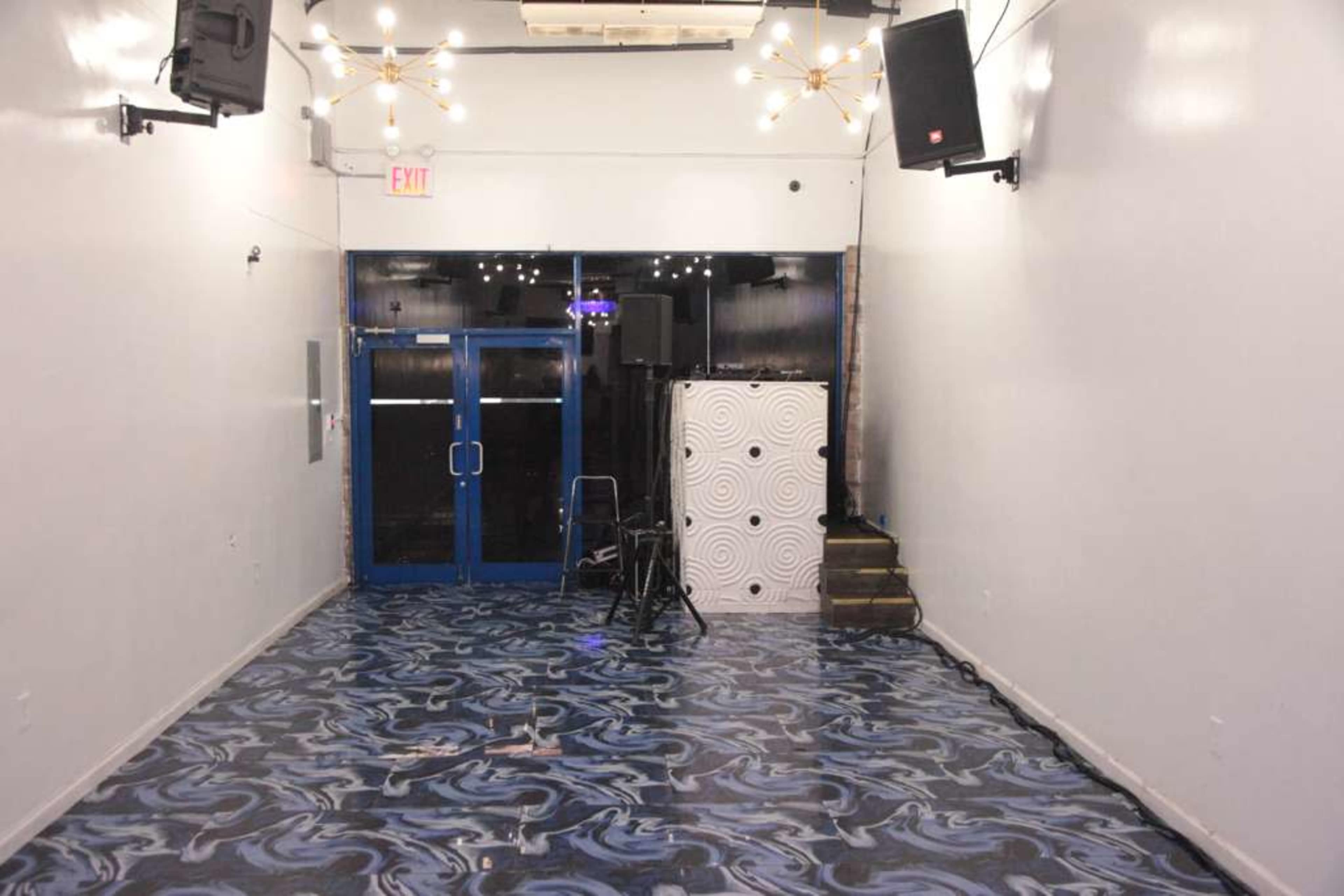 The image shows an empty room with a blue and black patterned floor, two large speakers mounted on the wall, and an entrance featuring double doors.