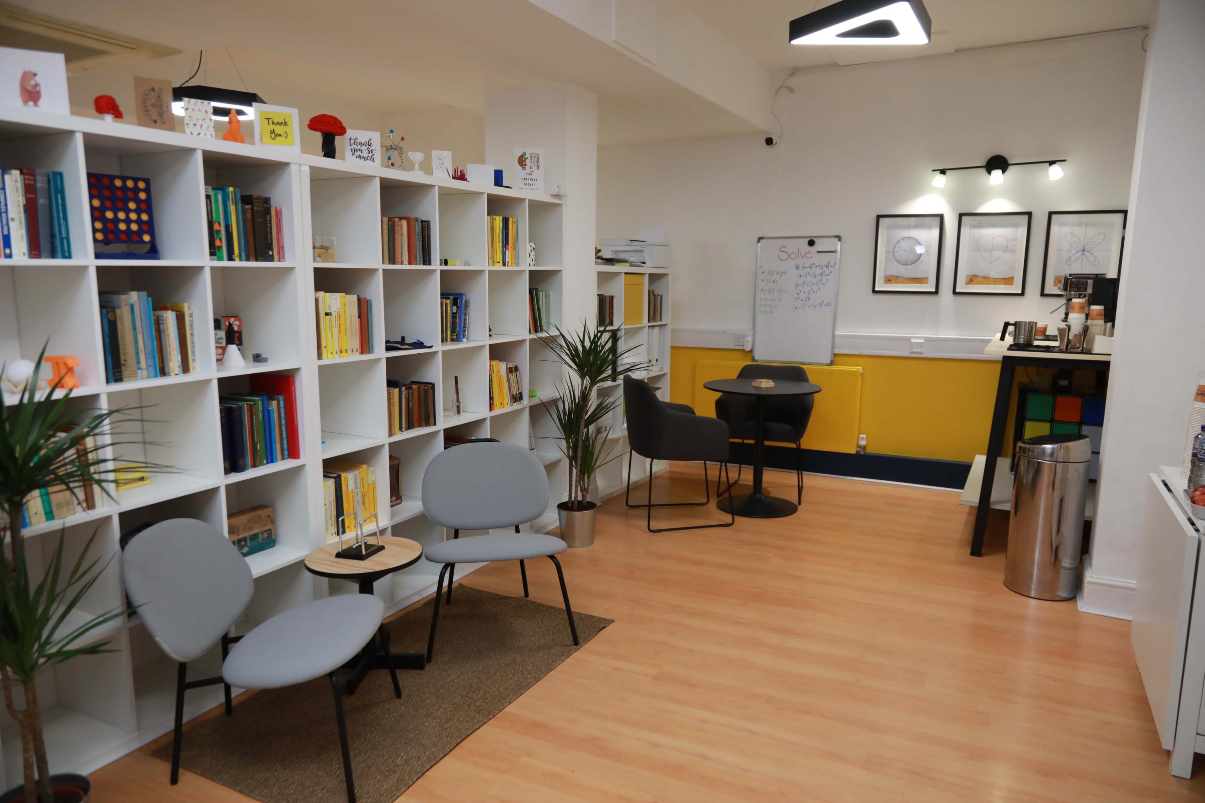 The image shows a well-lit modern reading room with bookshelves, a small table and chairs, and a whiteboard on the wall.