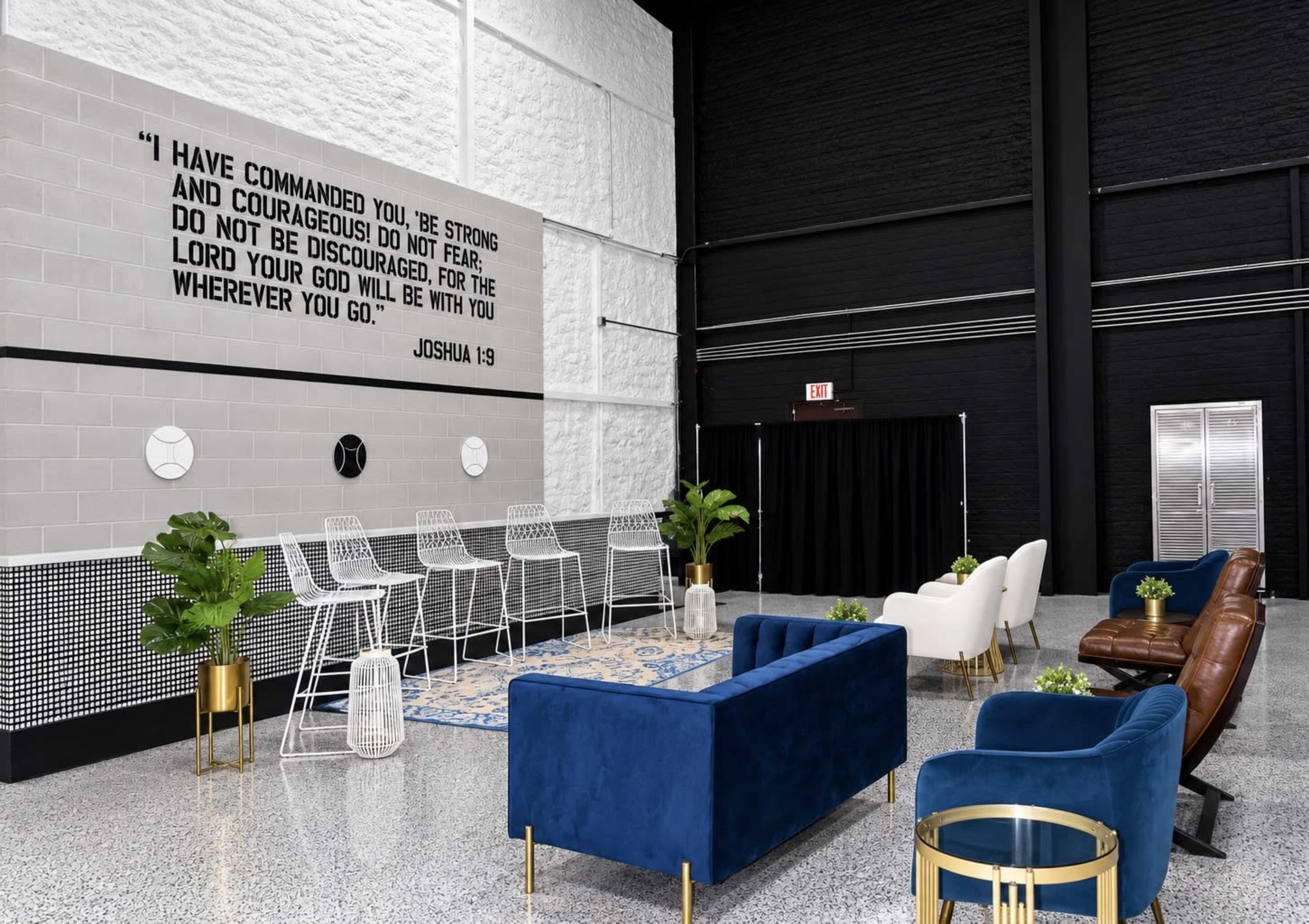The image shows a modern lounge area with a large wall featuring a scriptural quote, comfortable seating options, and decorative plants.