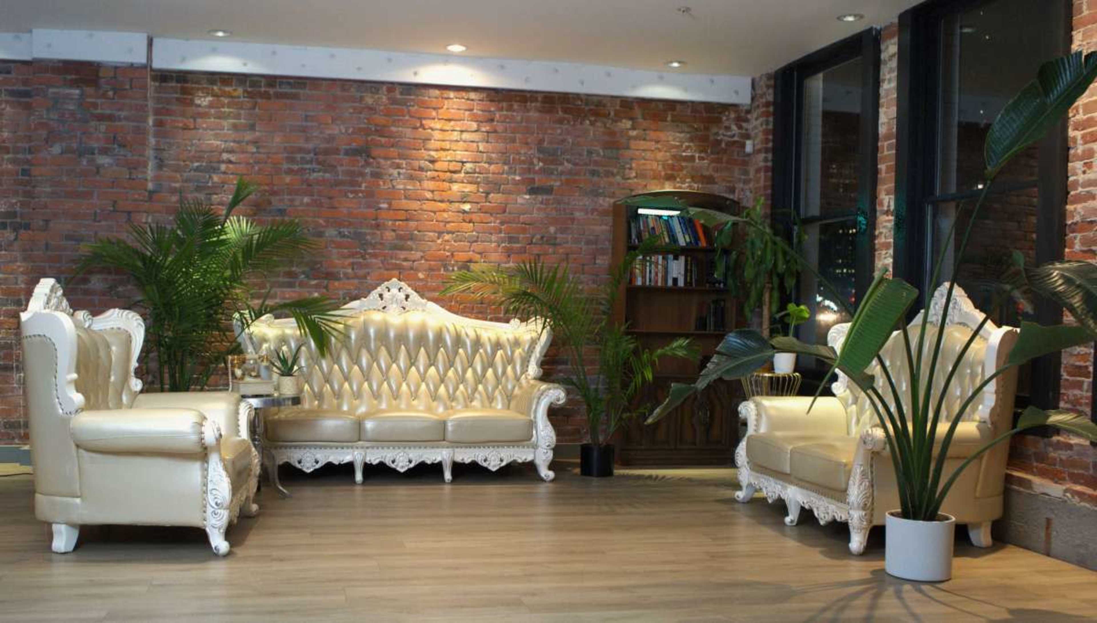 The scene features a traditional-style seating area with a cream-colored sofa and armchairs against a backdrop of exposed brick walls and potted plants.