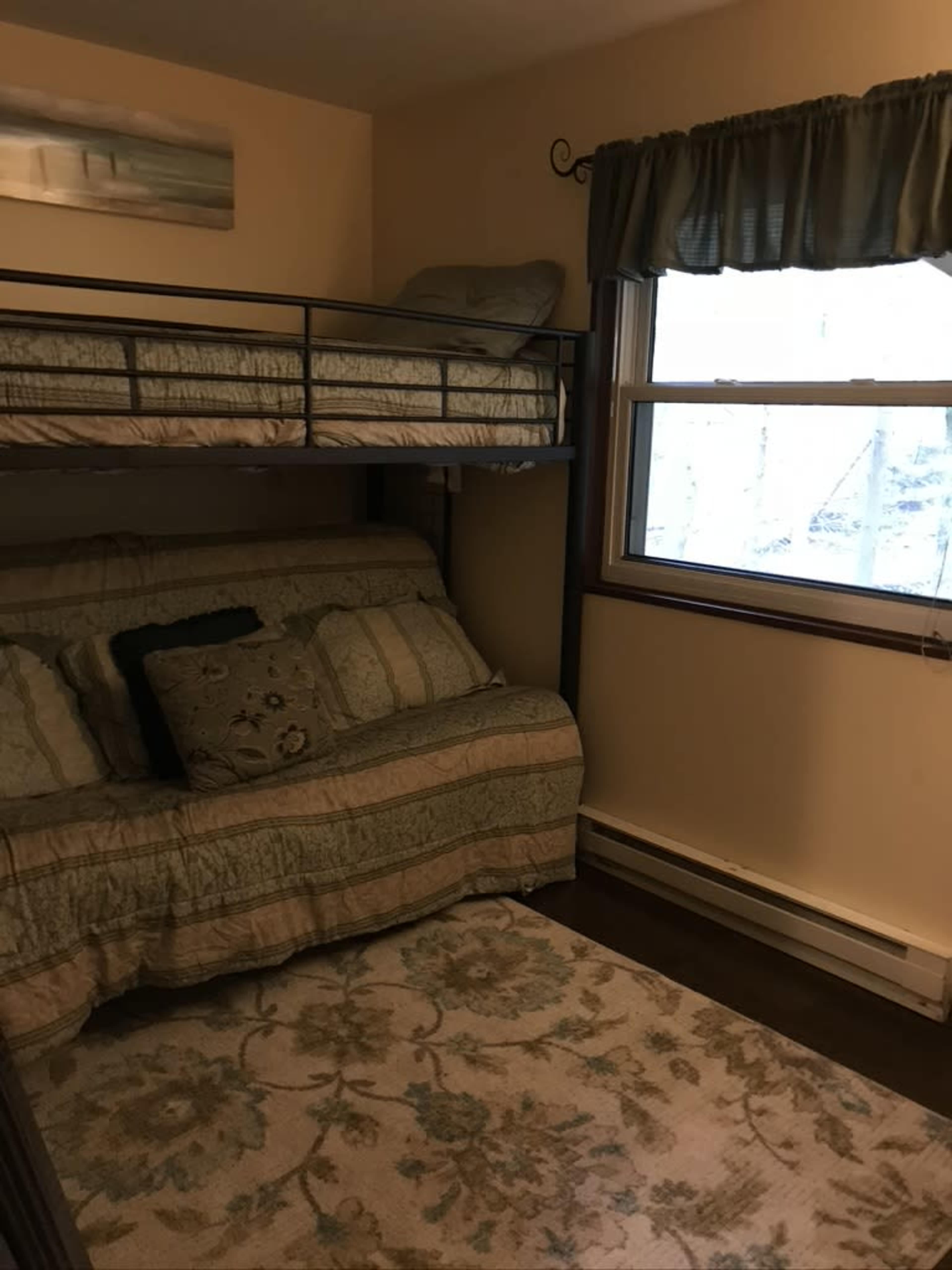 A small bedroom with a bunk bed, a sofa bed, and a window covered by a cream-colored curtain.