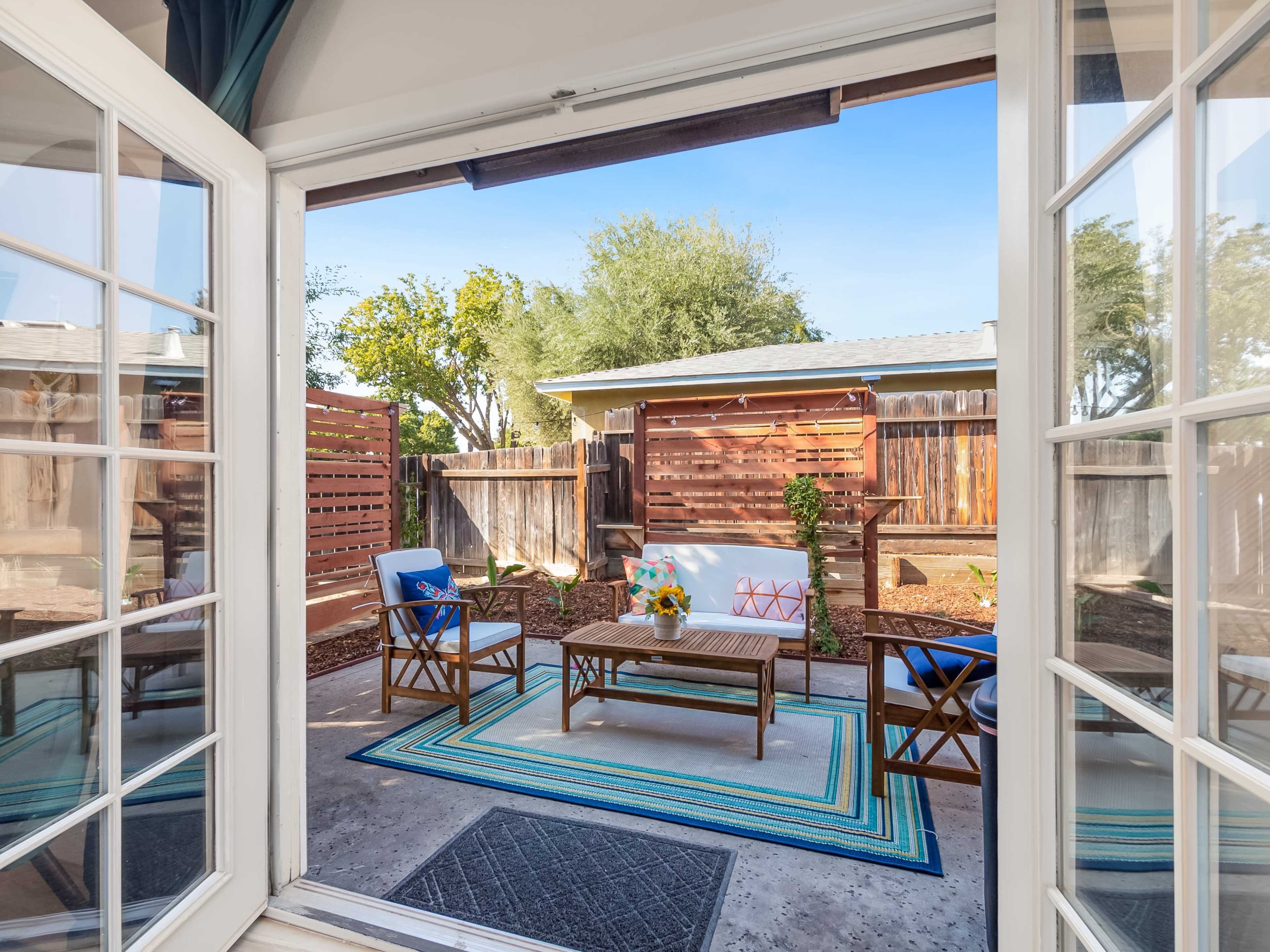A view through double doors reveals a patio area with a seating arrangement, colorful rug, and landscaped garden.