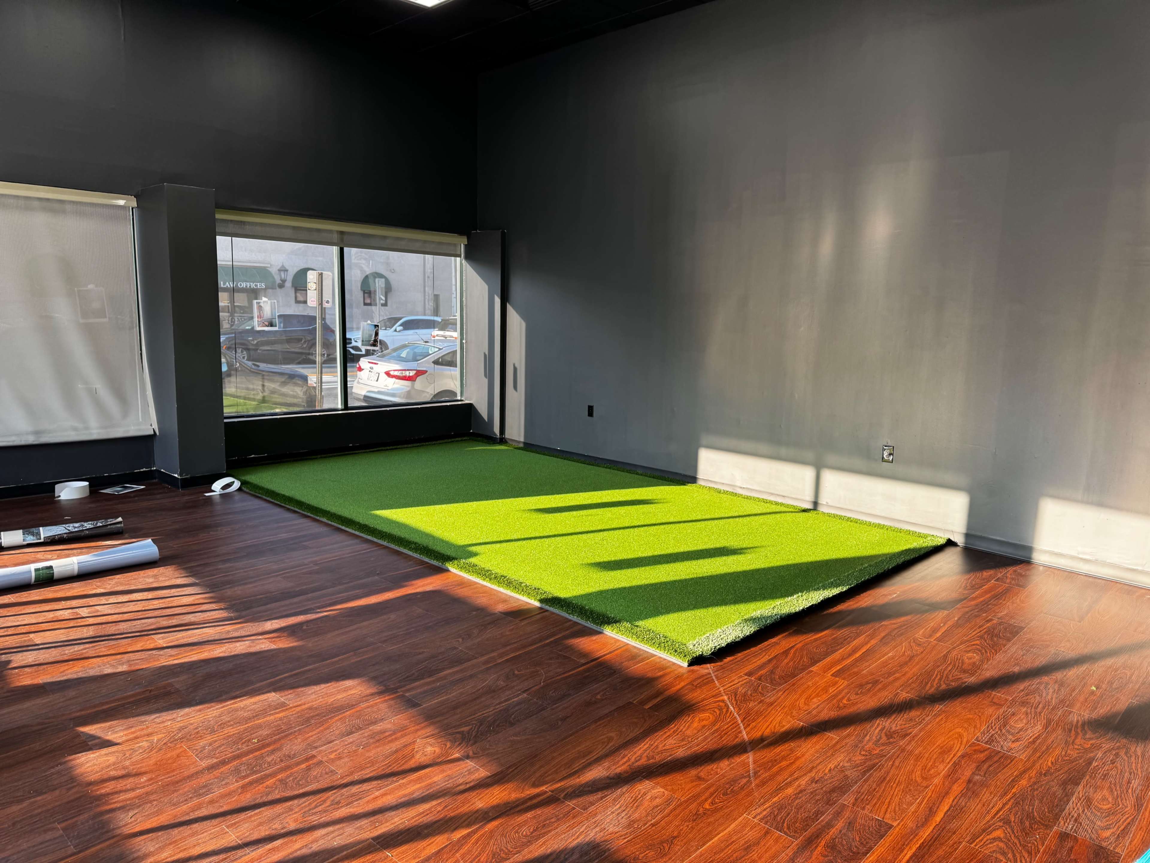 The image shows an empty room with a wooden floor, a rectangular piece of artificial green grass positioned against a window, and some rolled-up materials nearby.