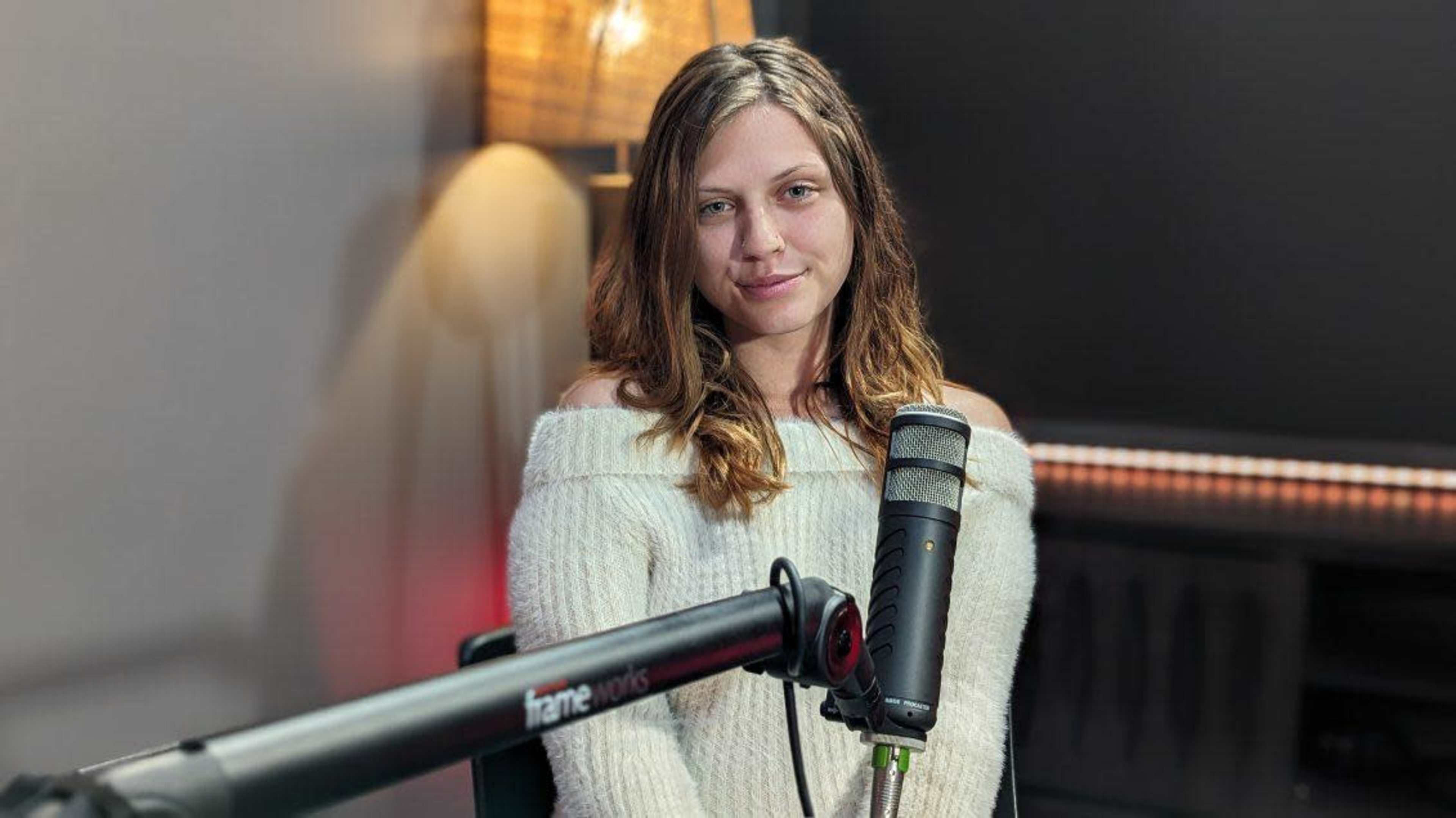 A person with long hair sits in front of a microphone in a softly lit room.