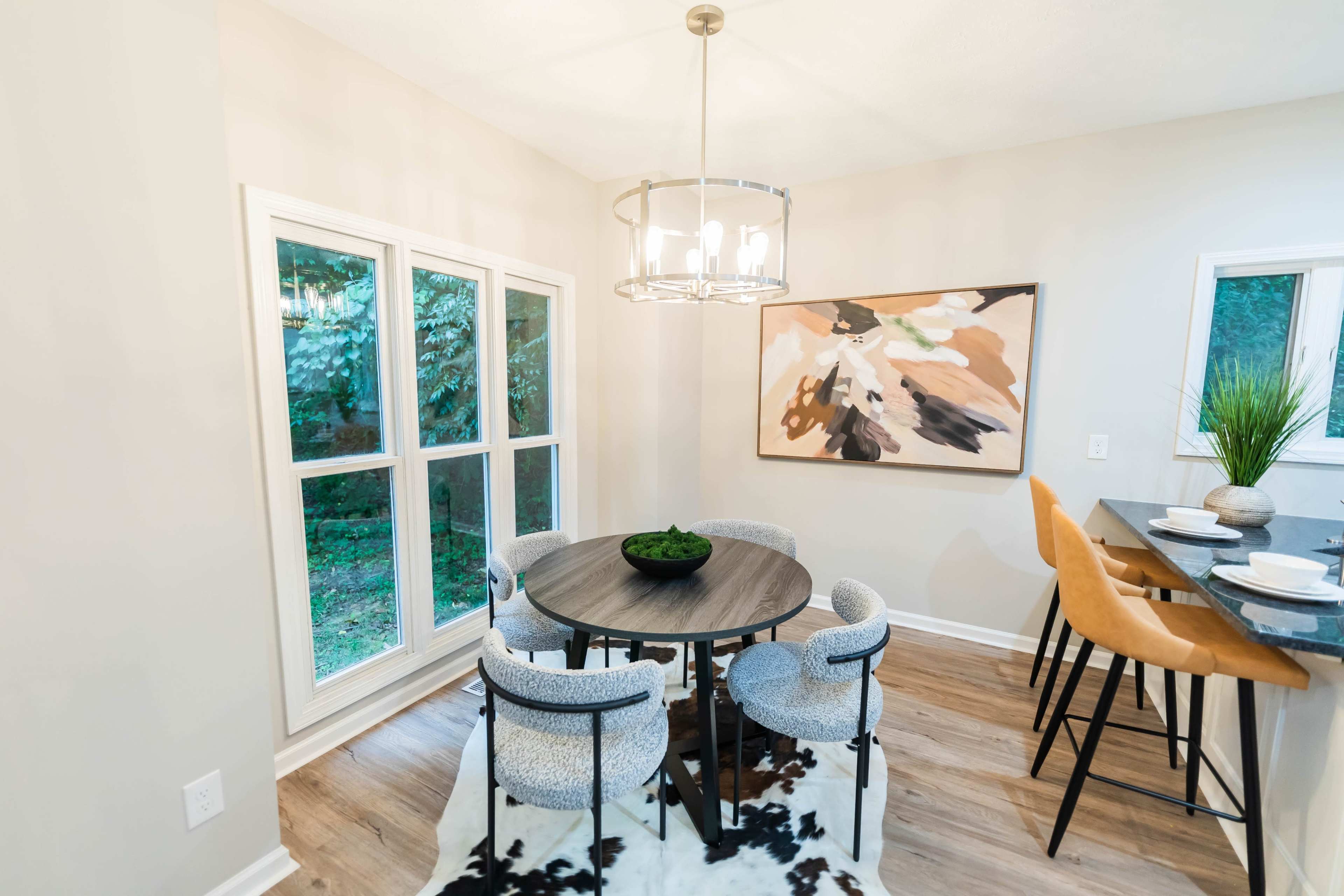 A modern dining area features a round table with upholstered chairs, a pendant light, and a large artwork on the wall, surrounded by large windows.