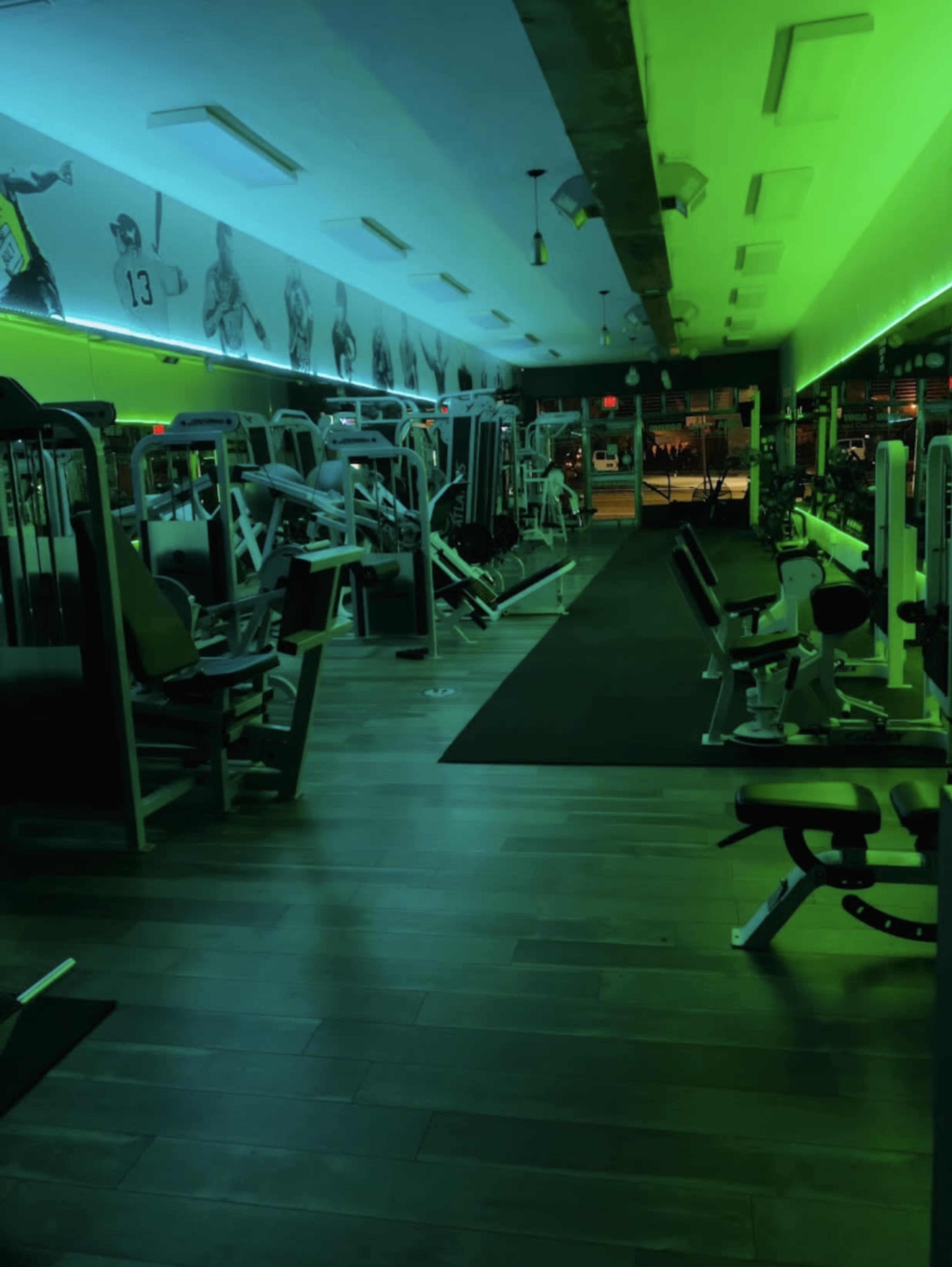 A dimly lit gym interior with fitness equipment lined up along the walls and a green ambient light.