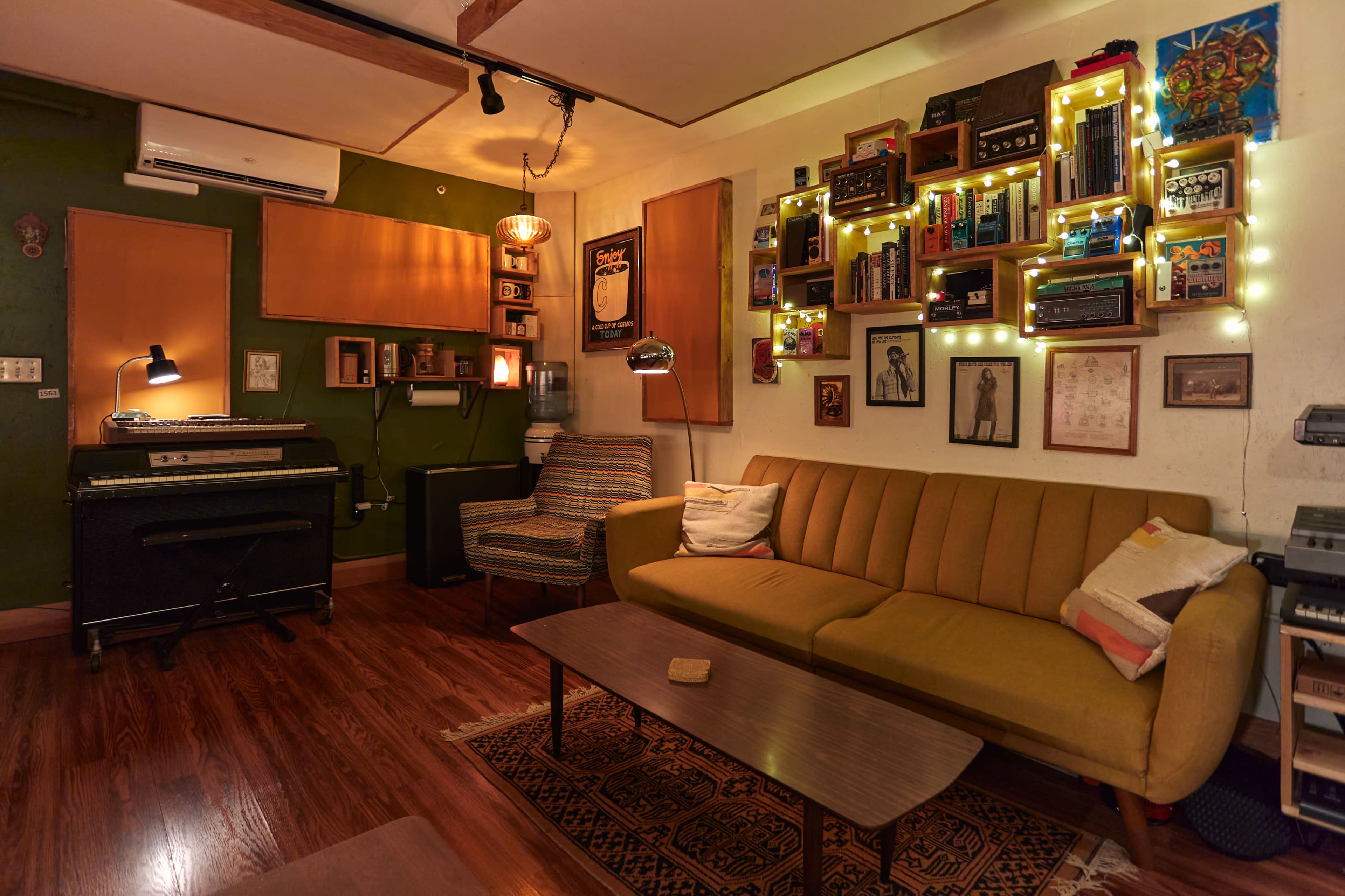 The image shows a cozy living room with a mustard-yellow sofa, a wooden coffee table, and various vintage music equipment displayed on the walls, illuminated by string lights.