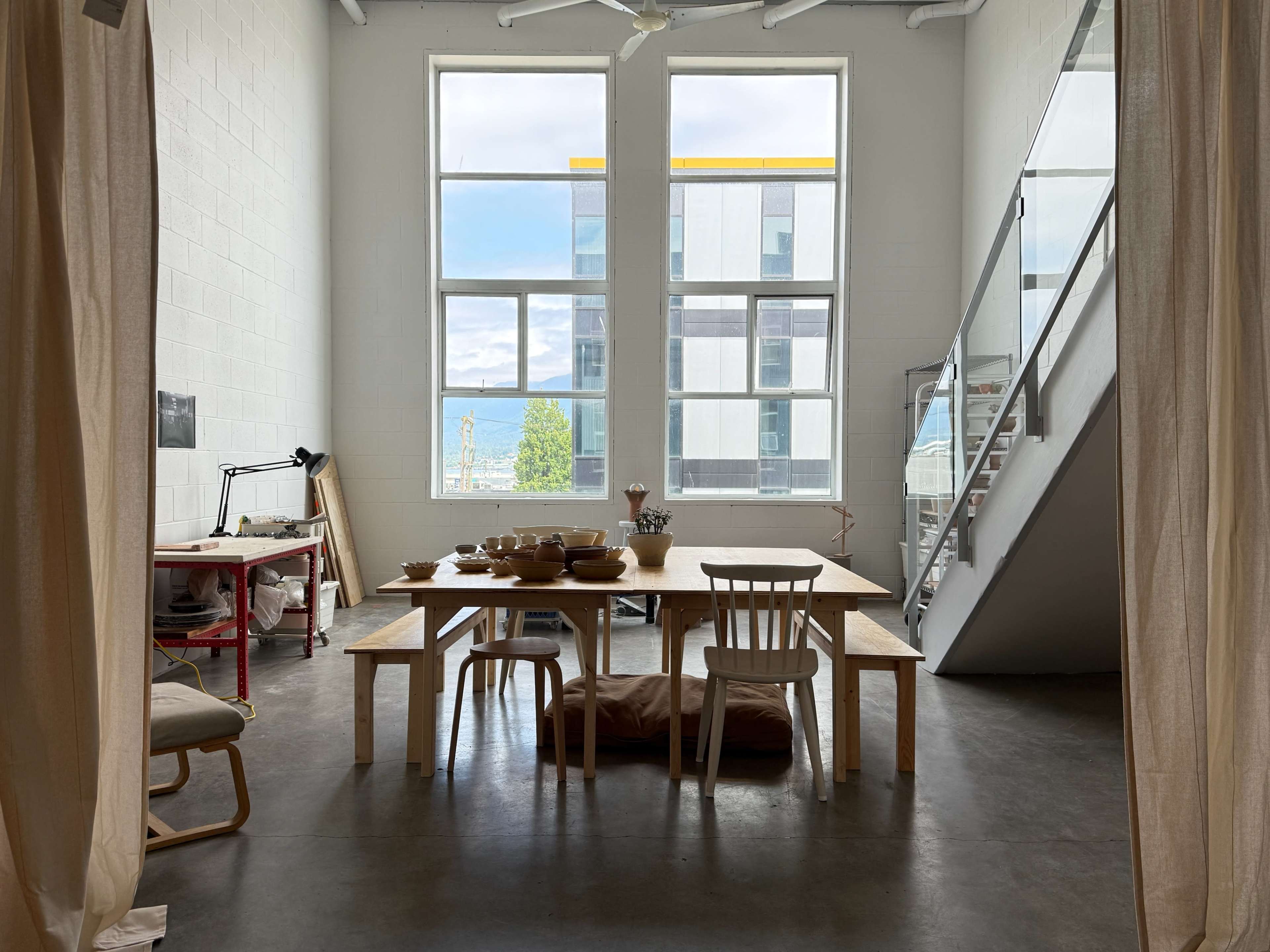 A bright, airy room features a large wooden dining table surrounded by chairs, with large windows allowing natural light and a view of an outdoor scene.