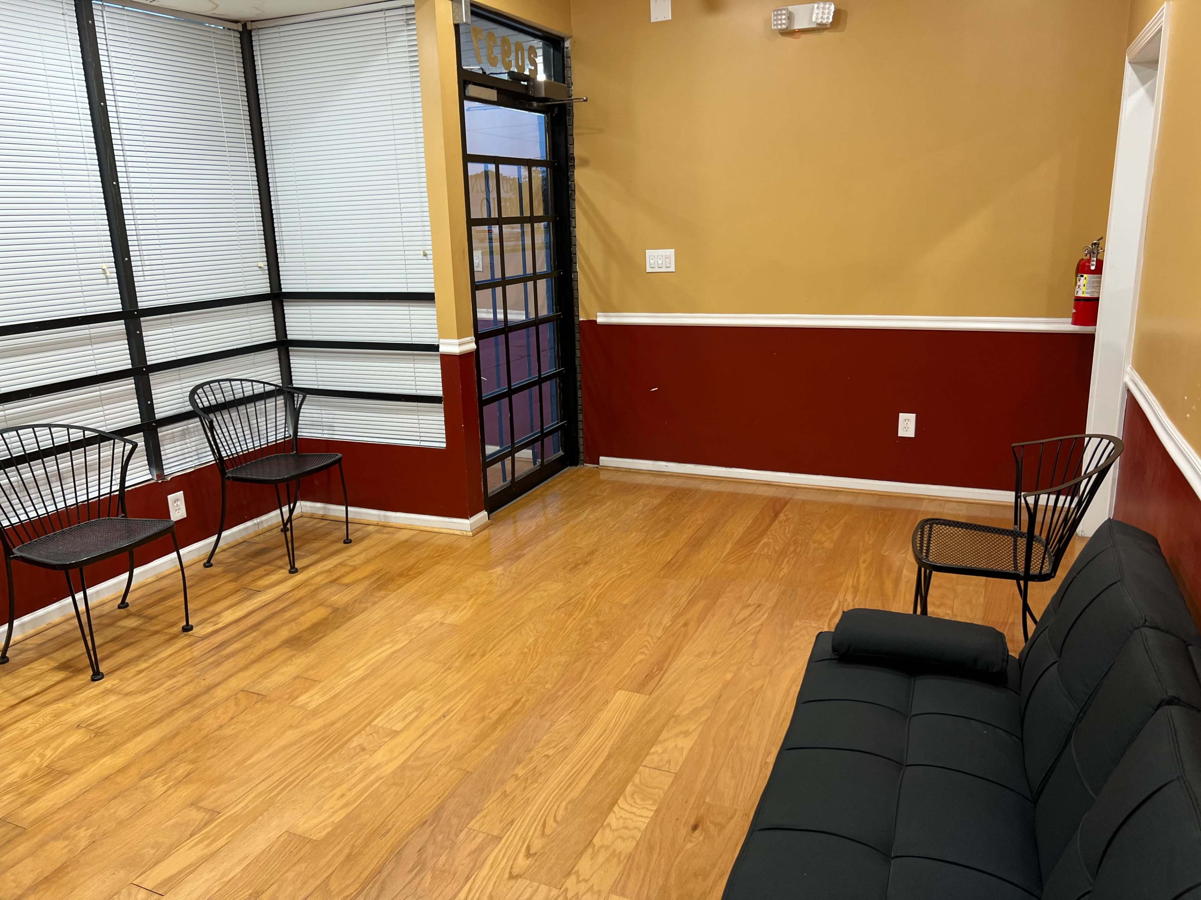 A small waiting area with a black sofa and a few metal chairs next to a door with large windows.