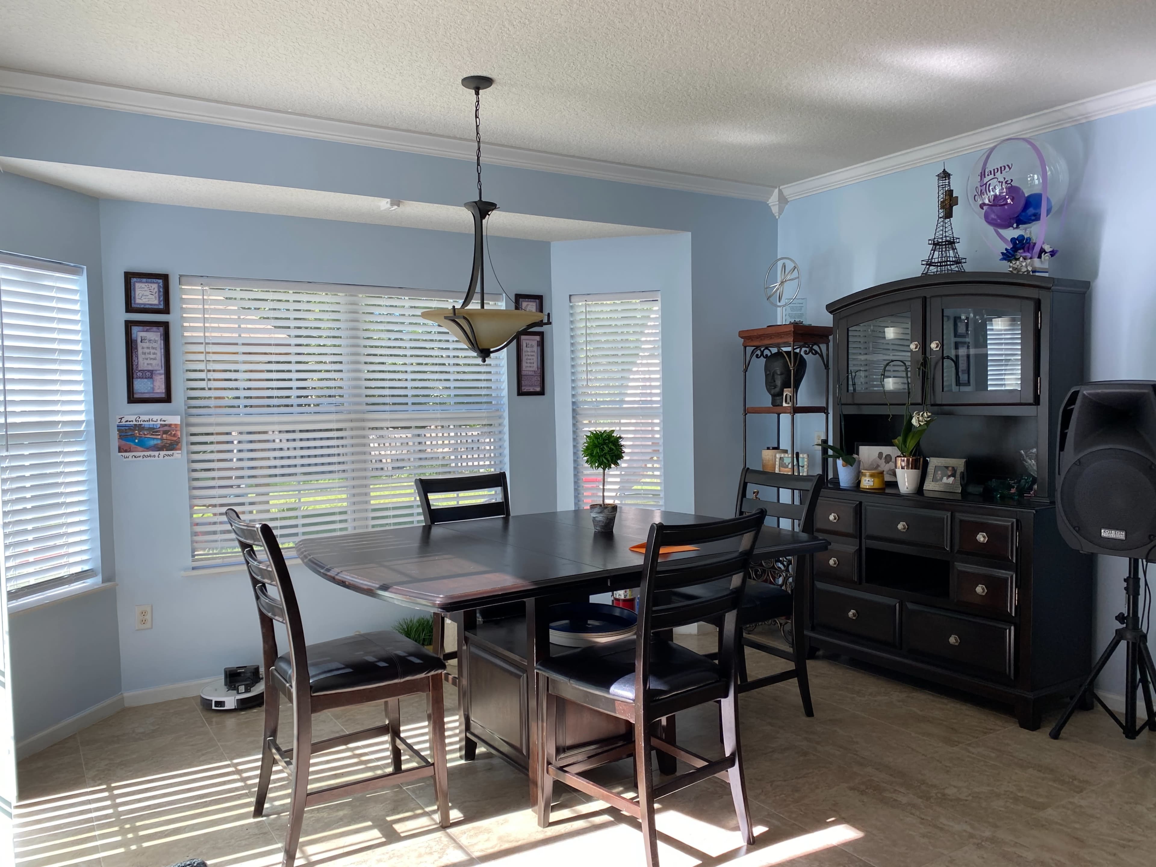 A dining area features a dark wooden table surrounded by chairs, with windows allowing natural light and a cabinet displaying decorative items.