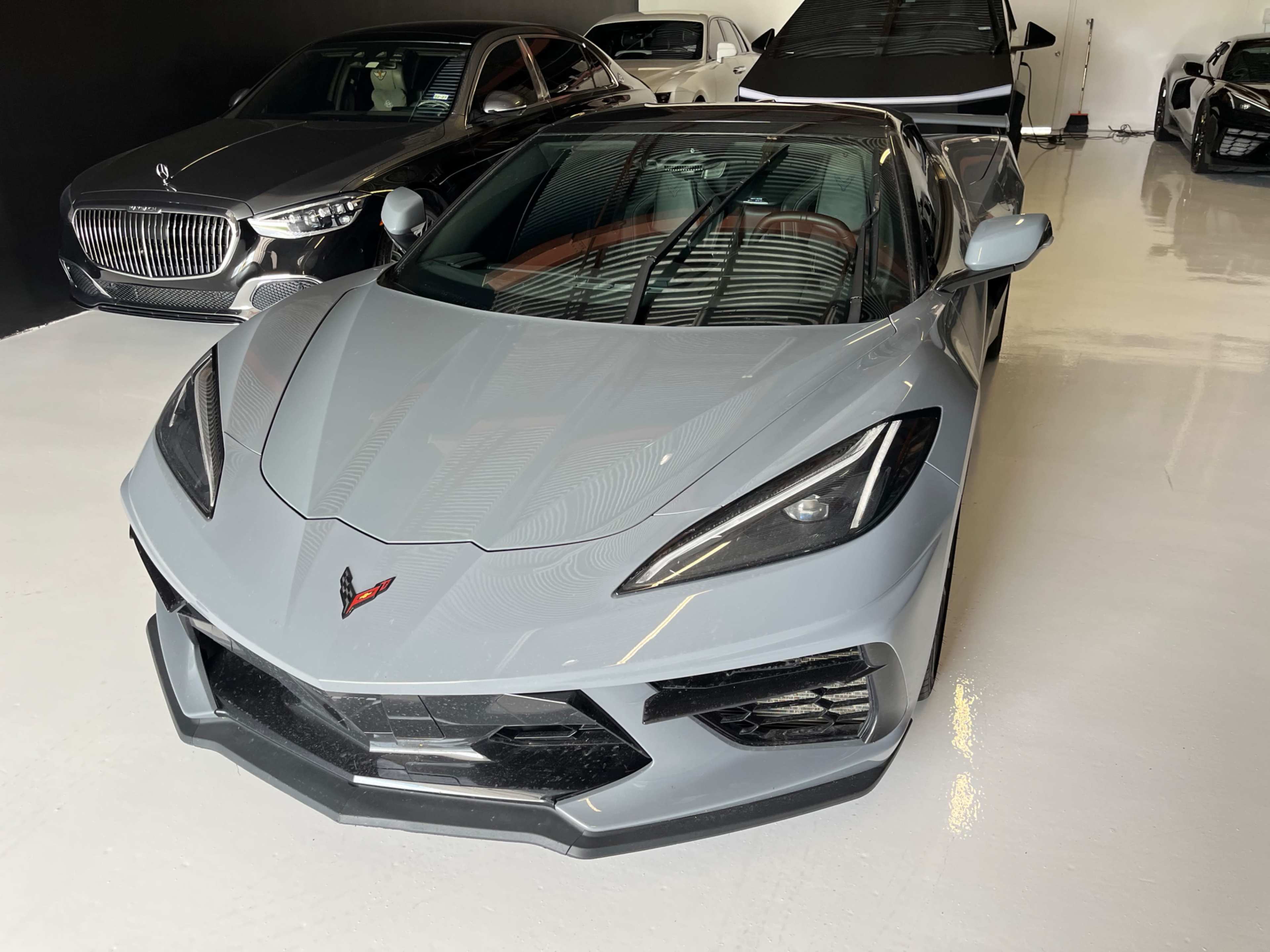 A gray sports car is parked in a garage featuring several luxury vehicles in the background.