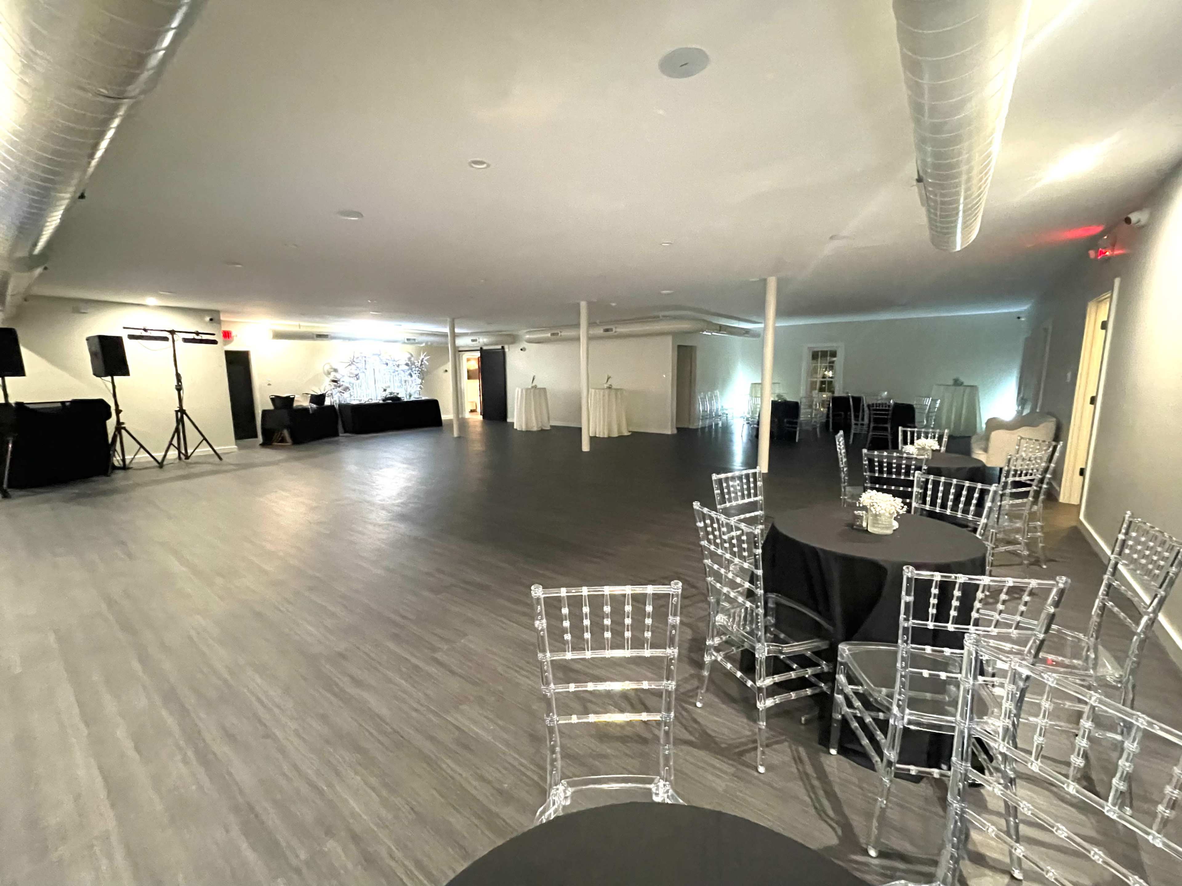 The image shows a spacious event venue featuring clear chairs and black tables, with a DJ setup in the corner and decorative drapes in the background.