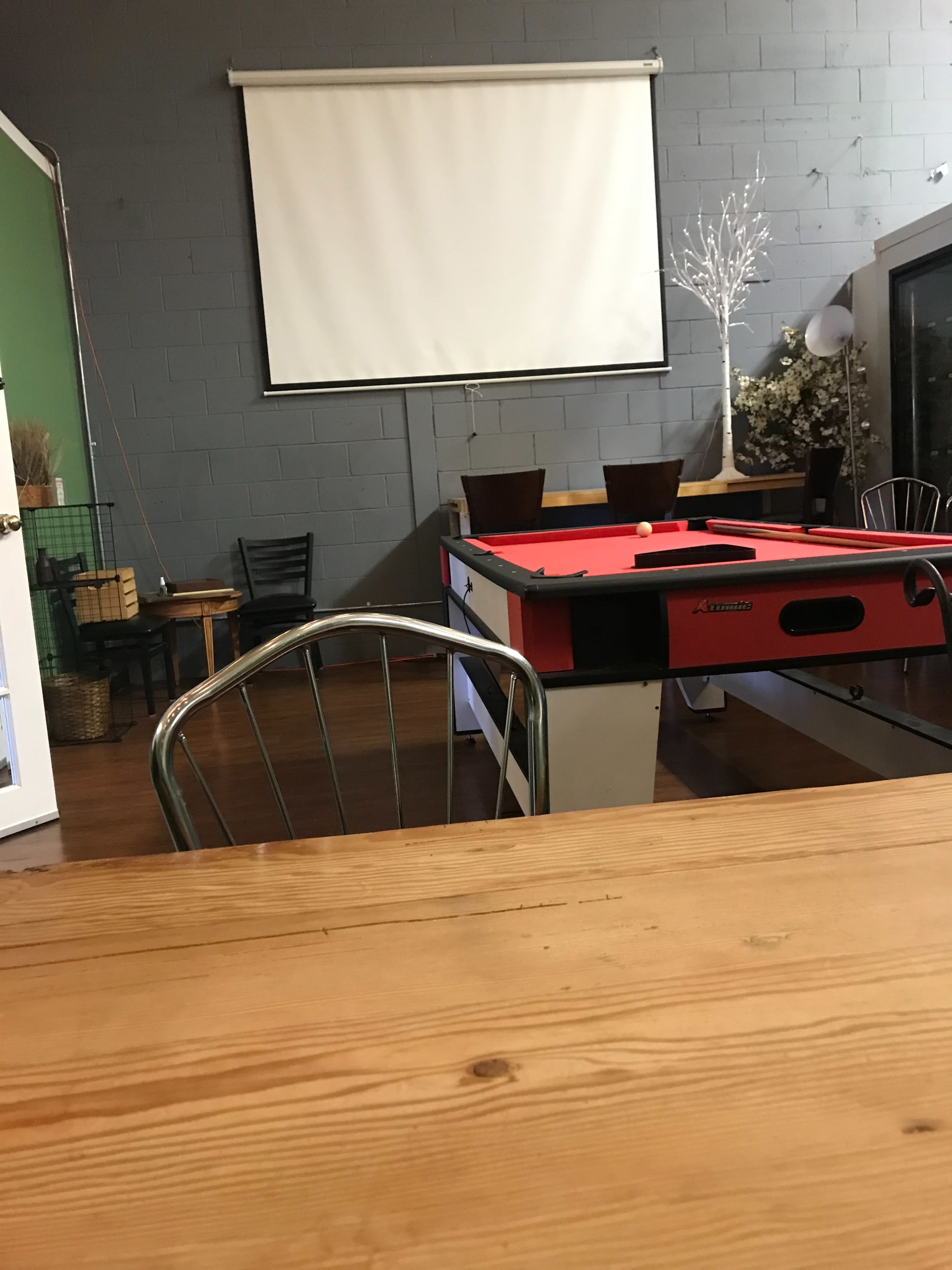 A wooden table with a metal chair in a room featuring a red air hockey table, a blank projection screen, and chairs against a gray wall.