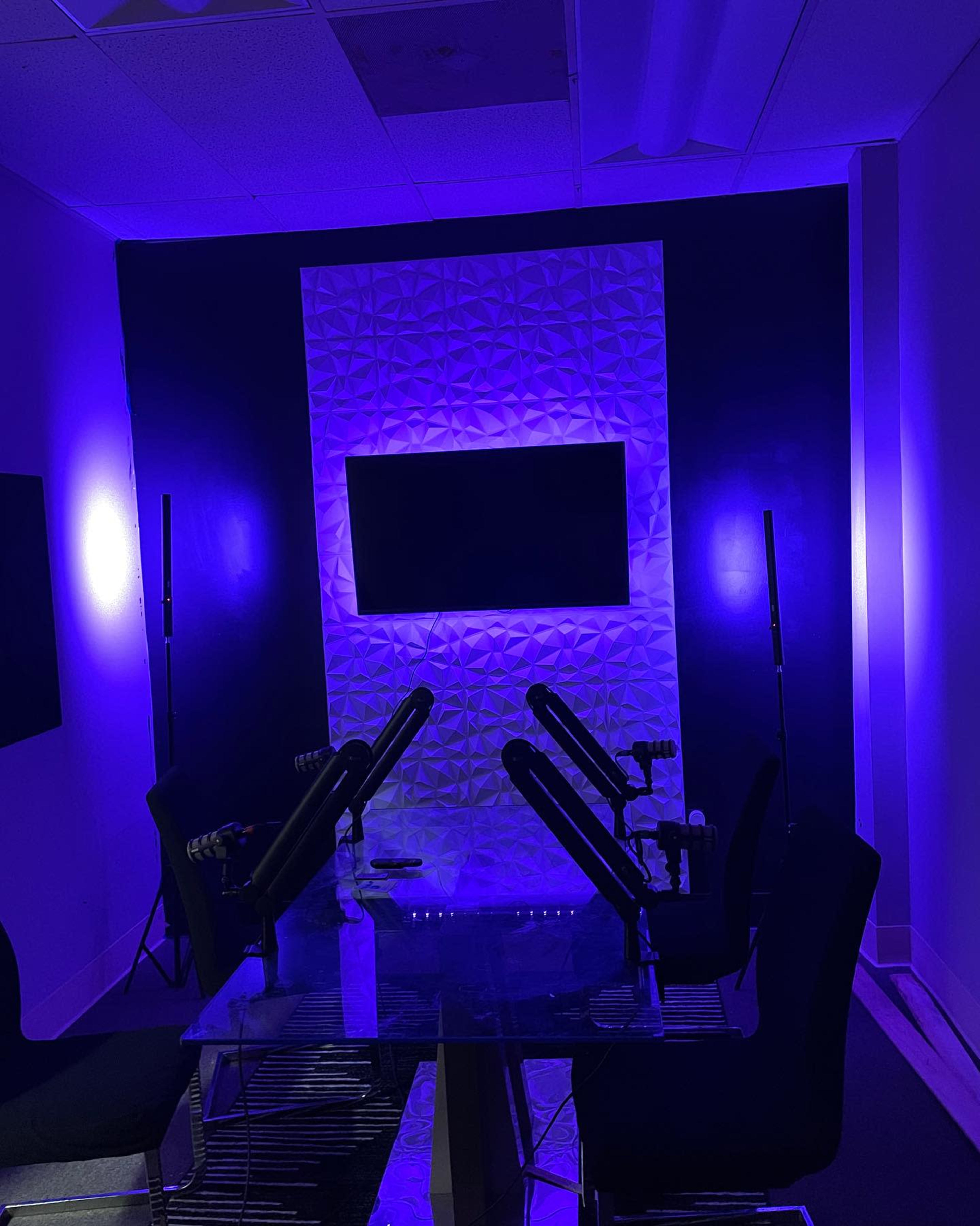 A podcasting room features a glossy table with microphones positioned for a discussion, surrounded by purple-lit walls and a television screen.