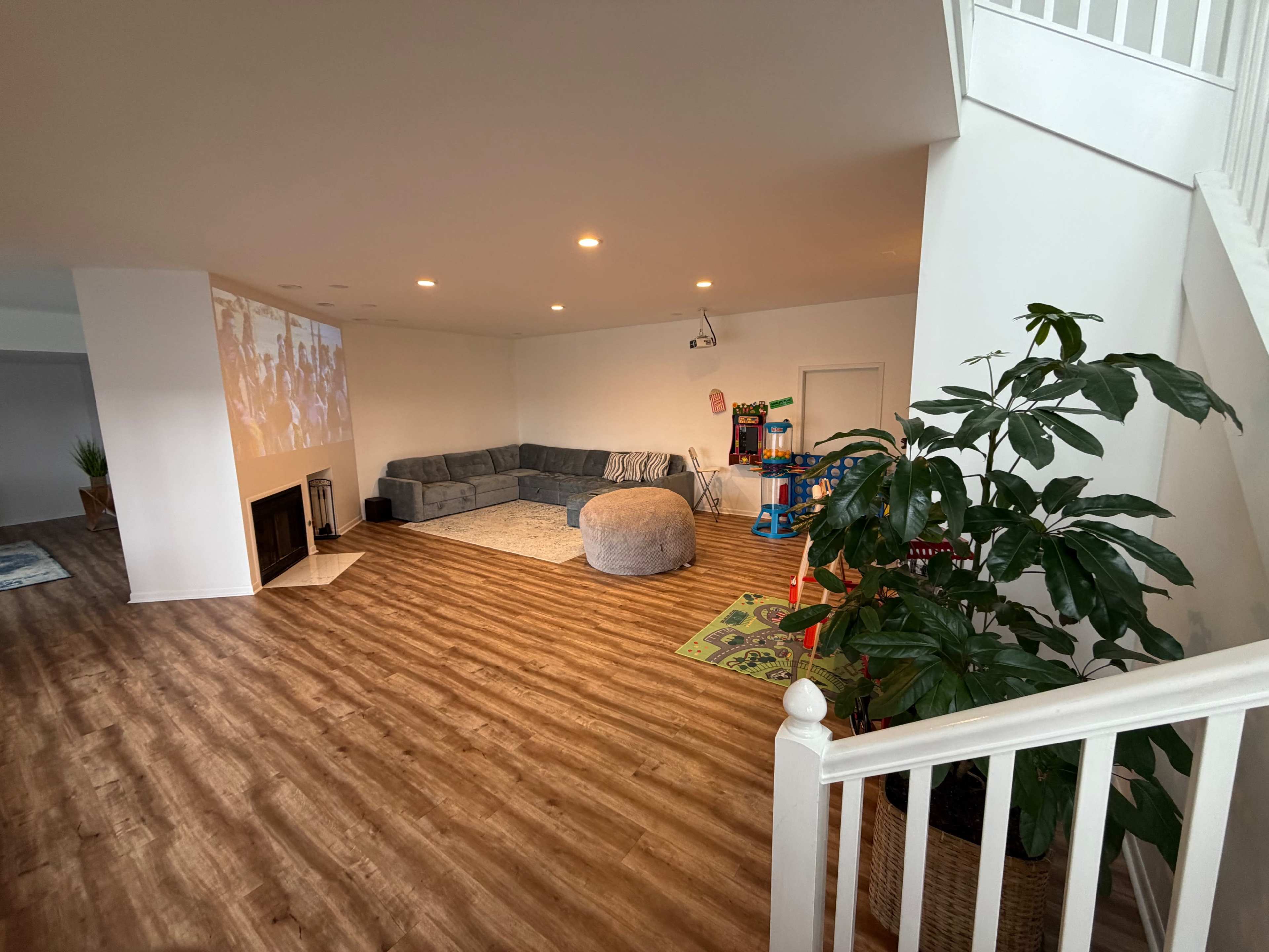The image shows a spacious living room with light-colored walls, a large gray sectional sofa, a round ottoman, a potted plant, and a play area with toys on a colorful mat.