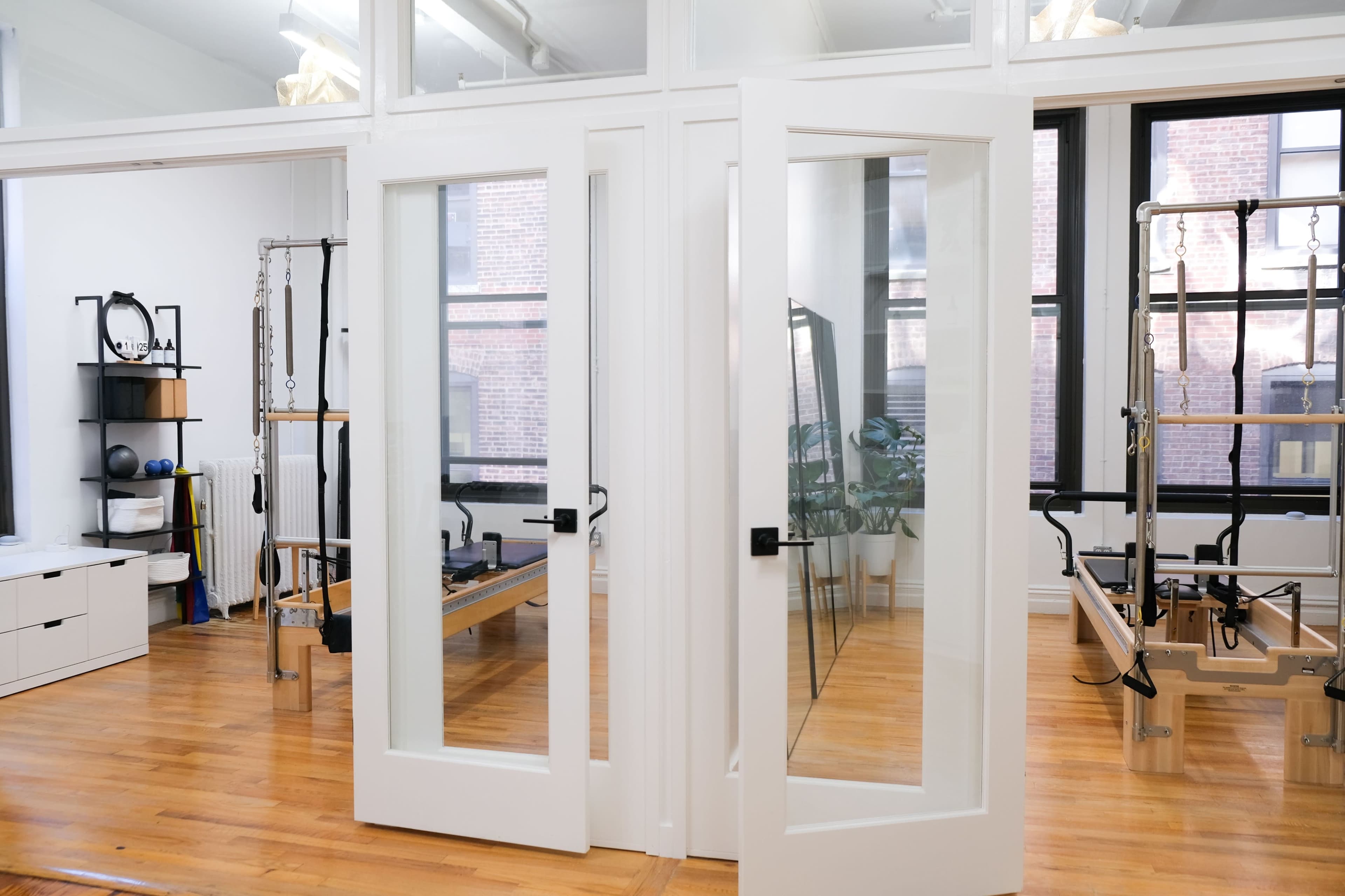 The image shows a well-lit exercise studio with wooden floors, featuring two glass-paneled doors that lead into separate areas equipped with fitness equipment.