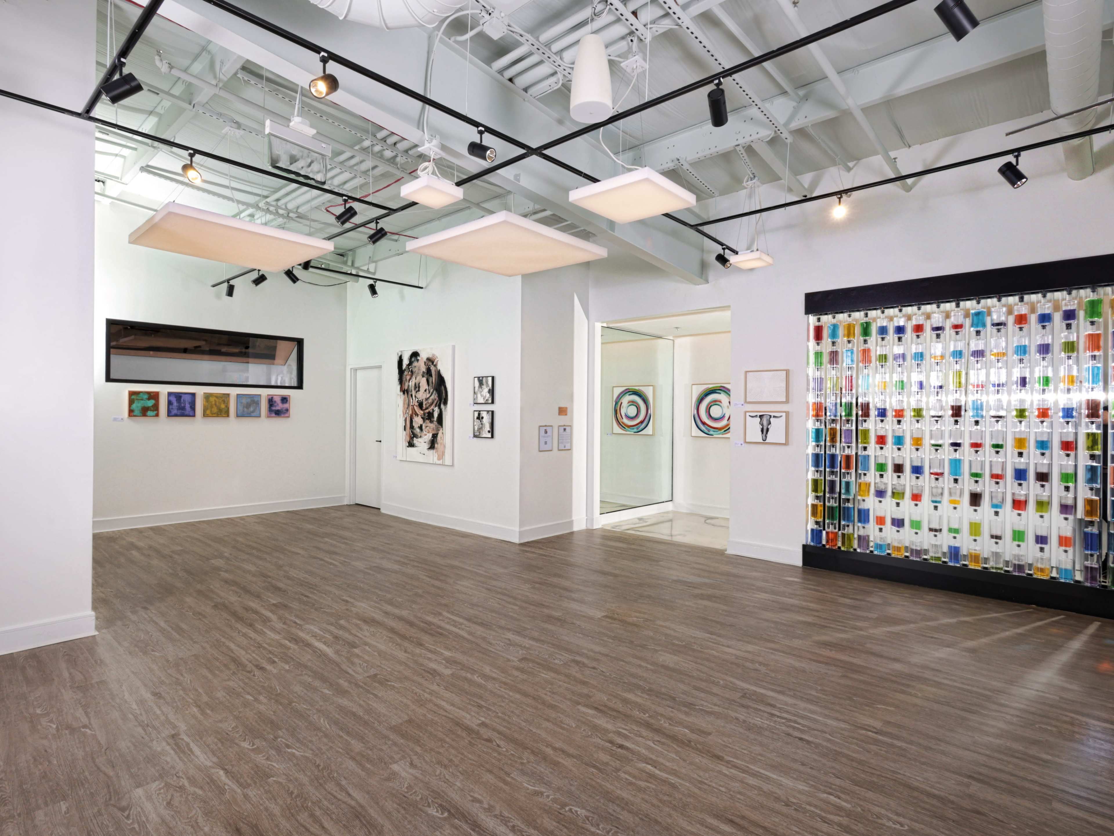 The image shows an art gallery with white walls, wooden flooring, and a variety of colorful artworks displayed on the walls.