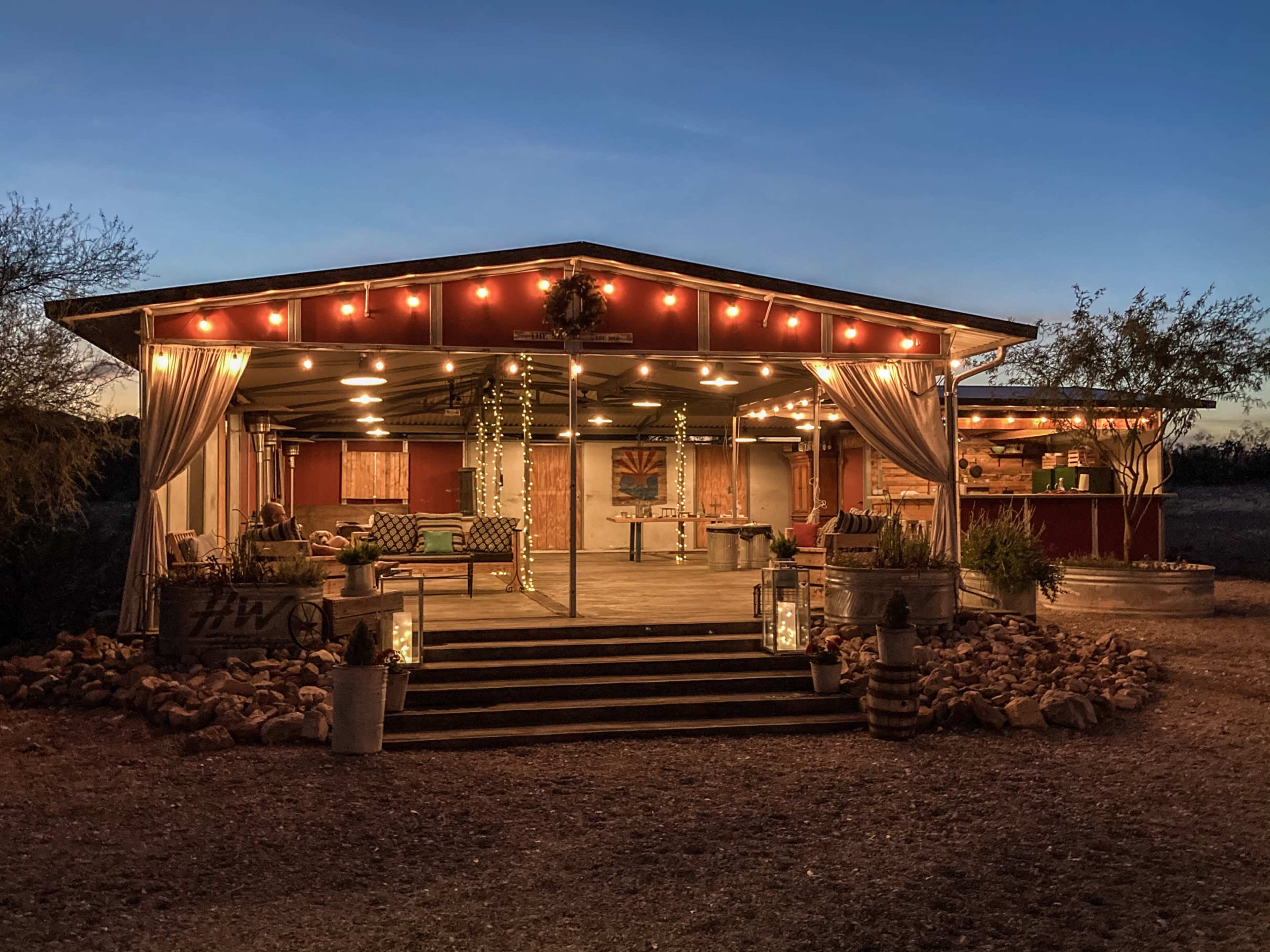 A rustic outdoor seating area with string lights and potted plants is set up in front of a wooden structure surrounded by gravel and desert vegetation.