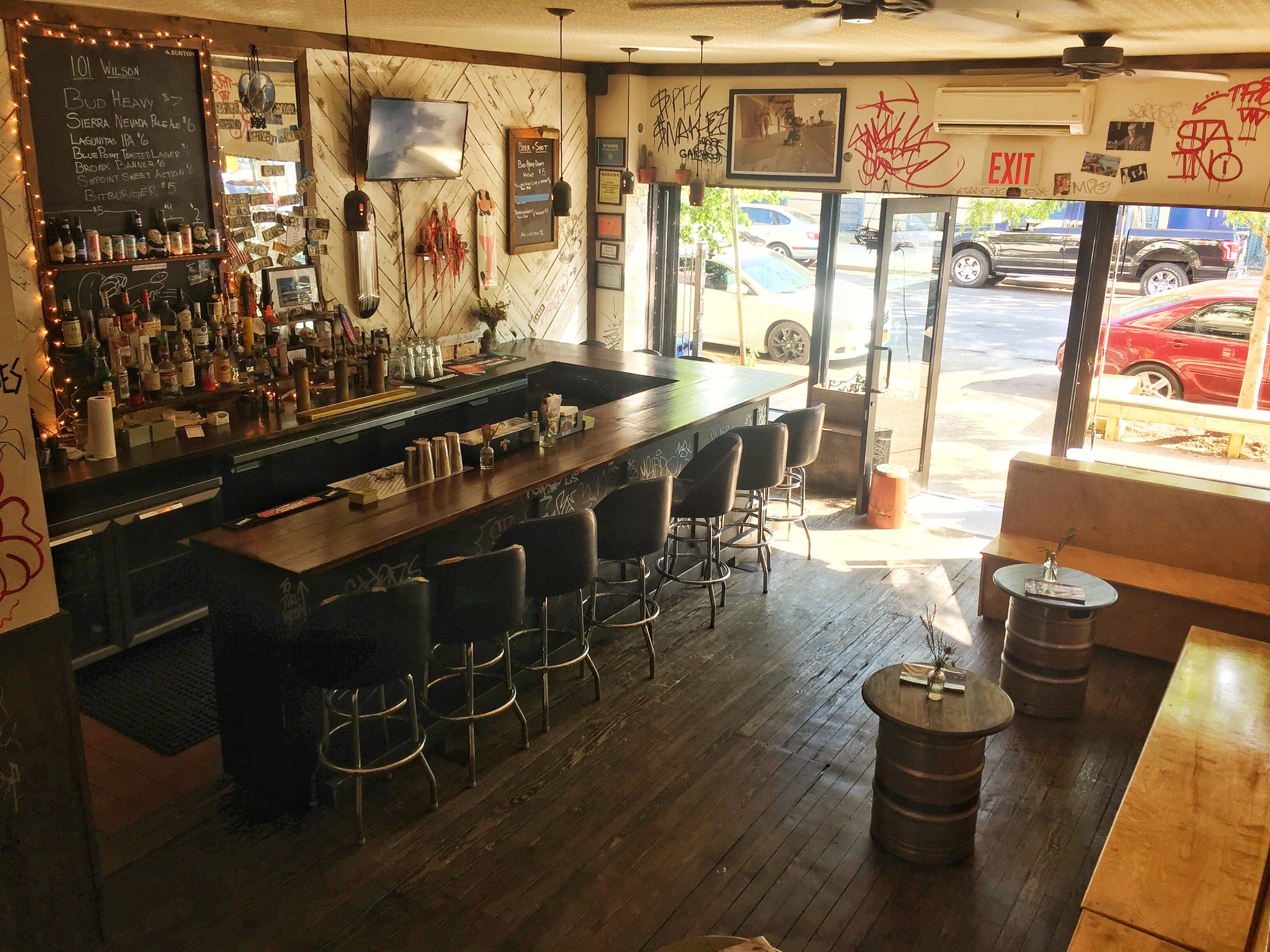The interior of a bar with a wooden counter, high stools, and large windows allowing light to enter.