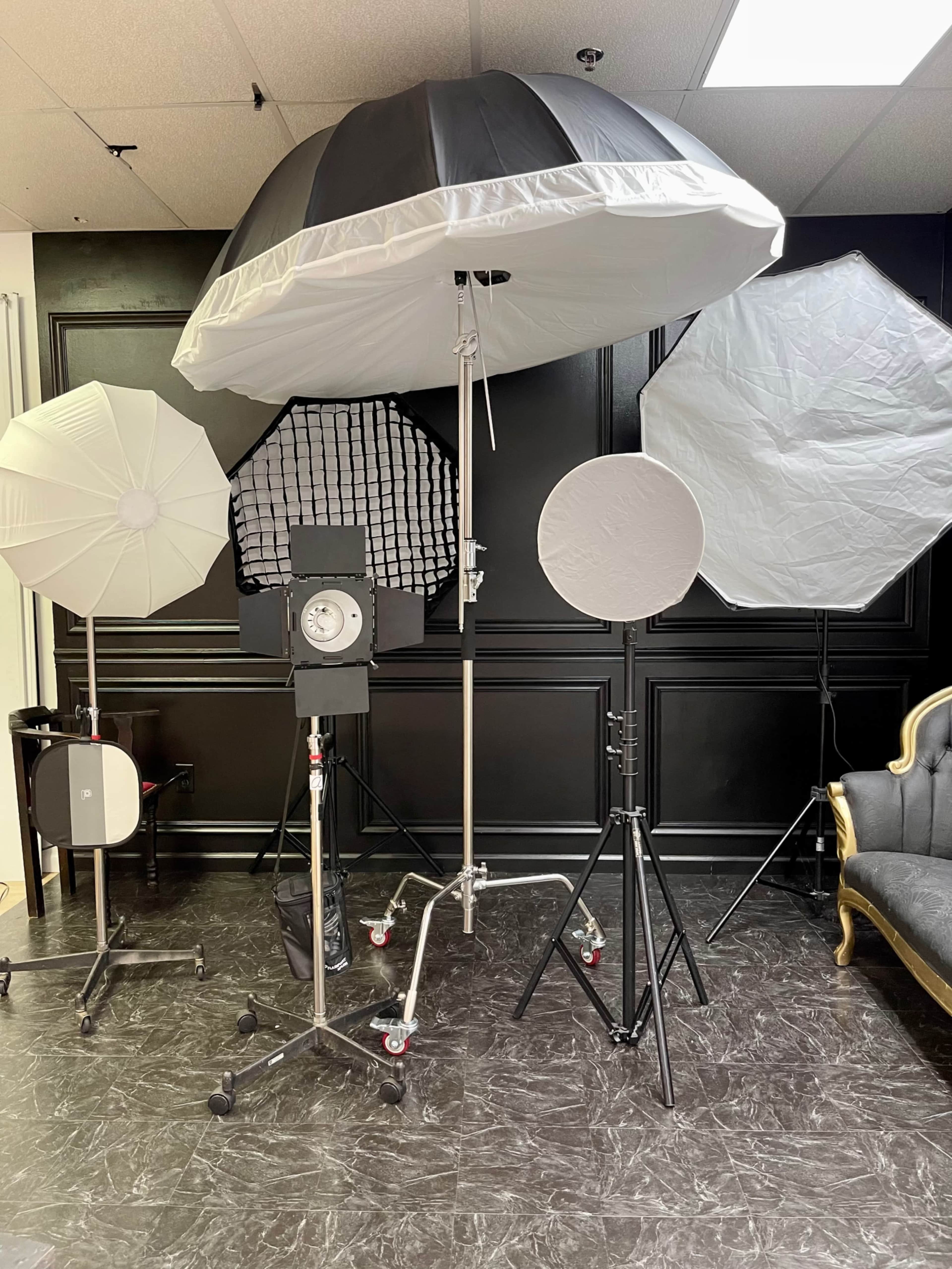 The image displays a photography studio setup featuring various light modifiers, including softboxes and umbrellas, arranged around a central light source.