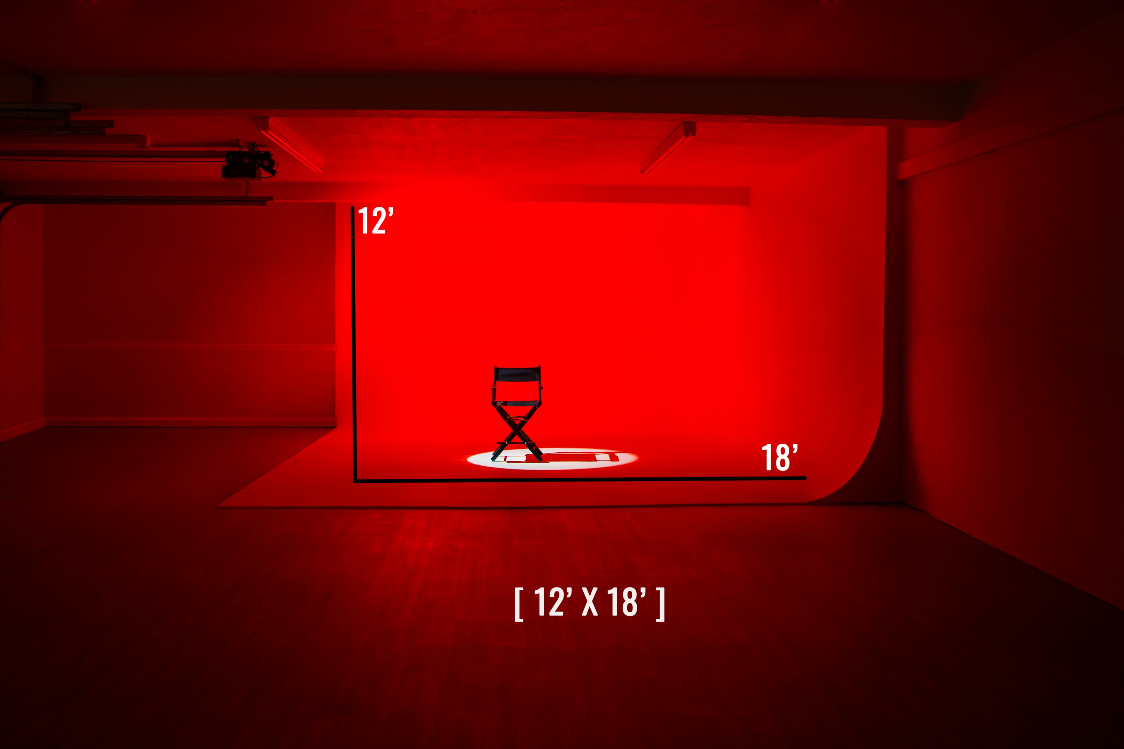 The image shows a dimly lit studio space featuring a black folding chair illuminated by a spotlight on a red background, with dimensions noted as 12 feet by 18 feet.