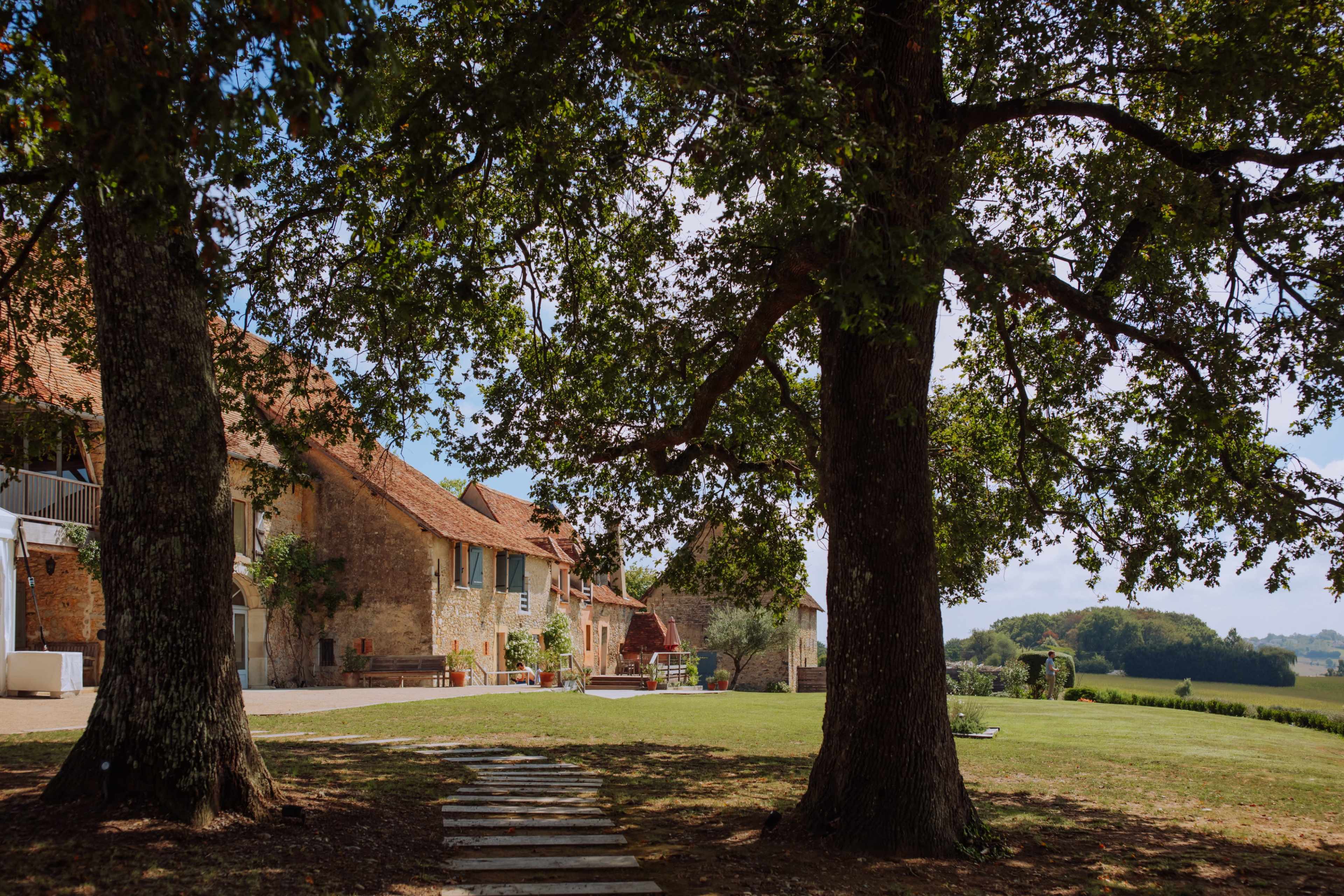 Two large trees frame a stone house with red-tiled roofs, surrounded by a grassy area and distant hills.
