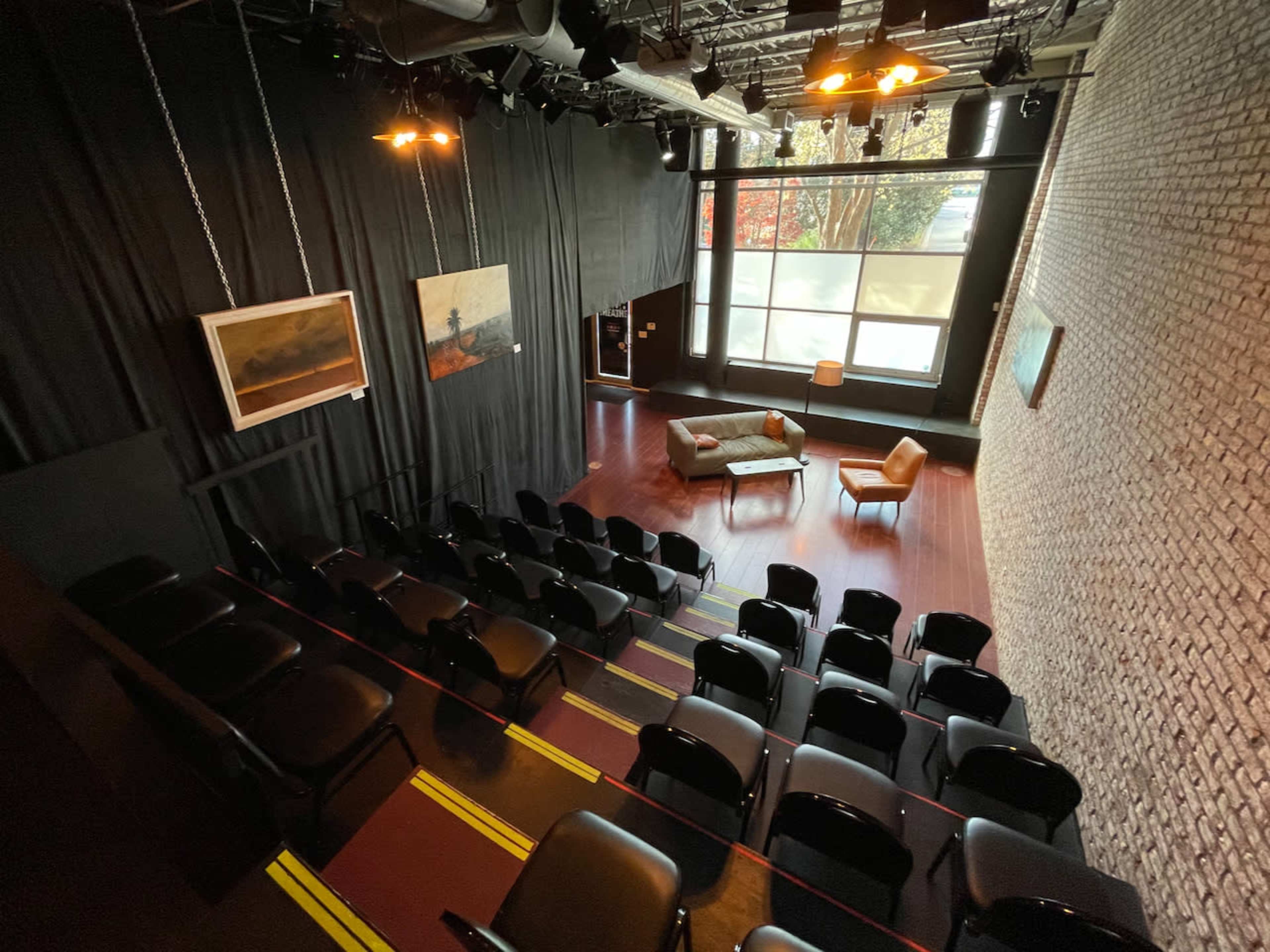 The image shows an auditorium-style space with rows of black chairs facing a small stage area that features a couch and a lamp, surrounded by large windows and artwork on the walls.