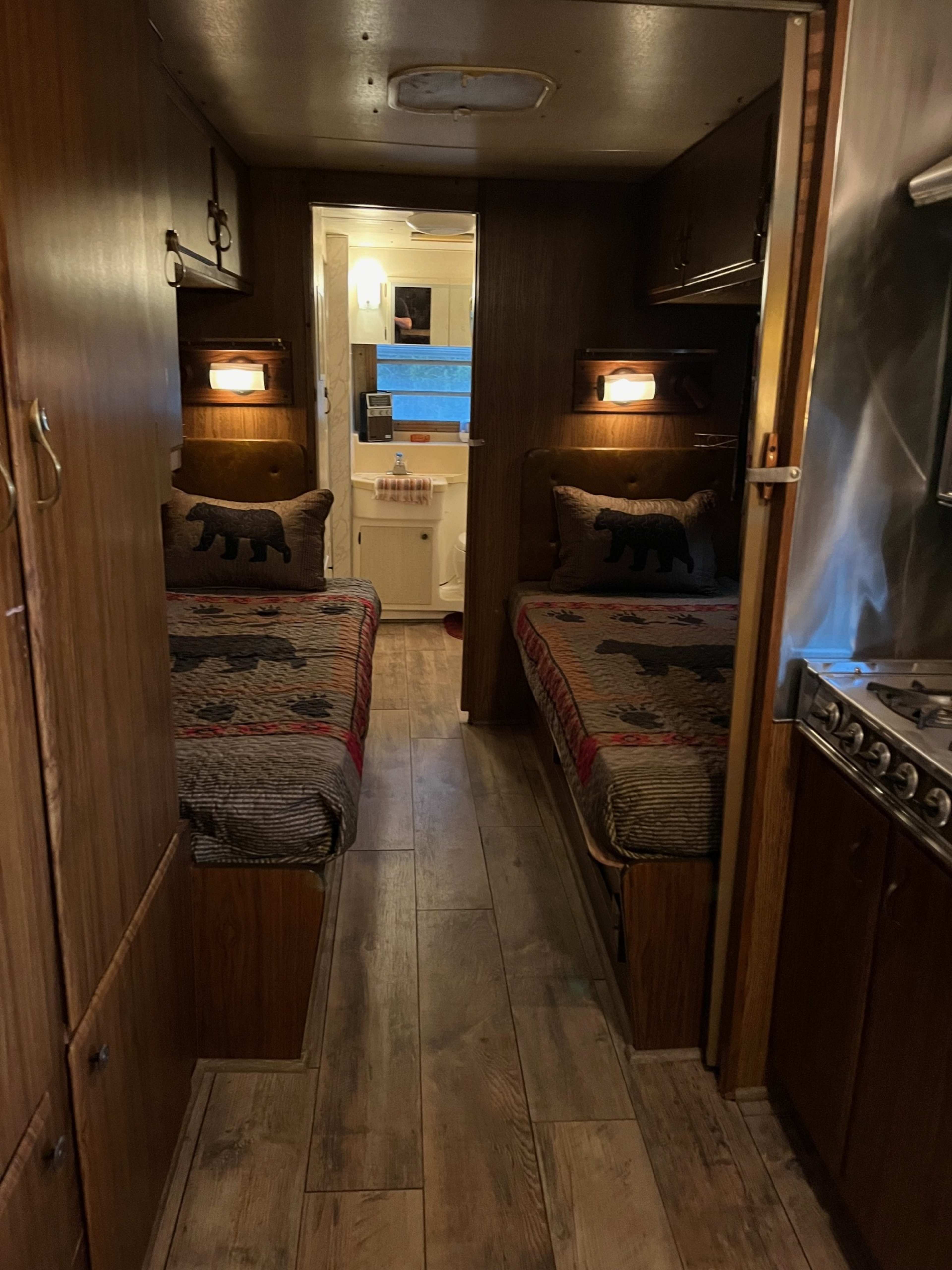 The interior of a vintage travel trailer features two twin beds with patterned blankets, a small kitchen area, and a bathroom at the far end.