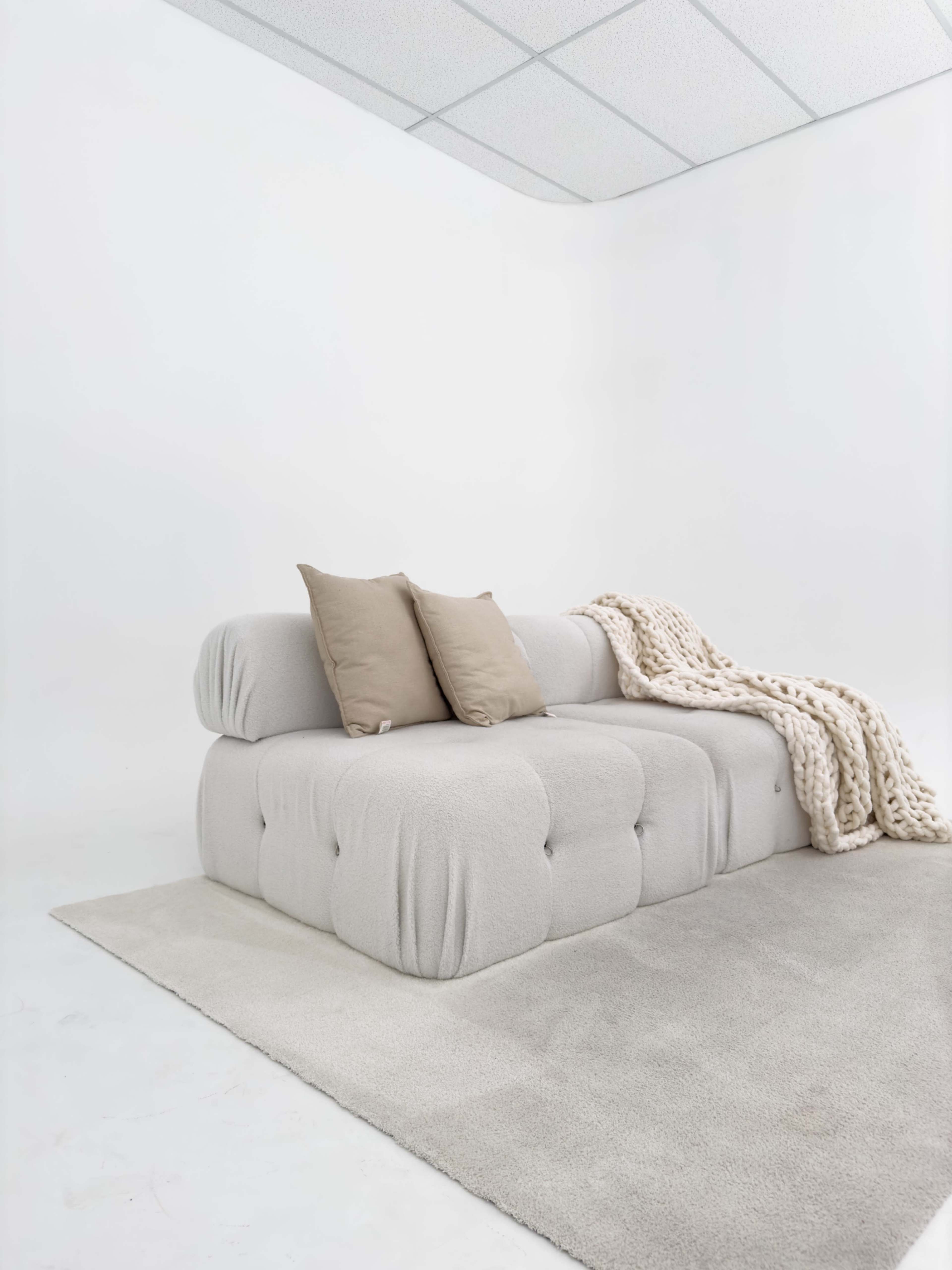 A light gray tufted sofa with two beige pillows and a beige knitted throw blanket sits on a light rug against a white wall.
