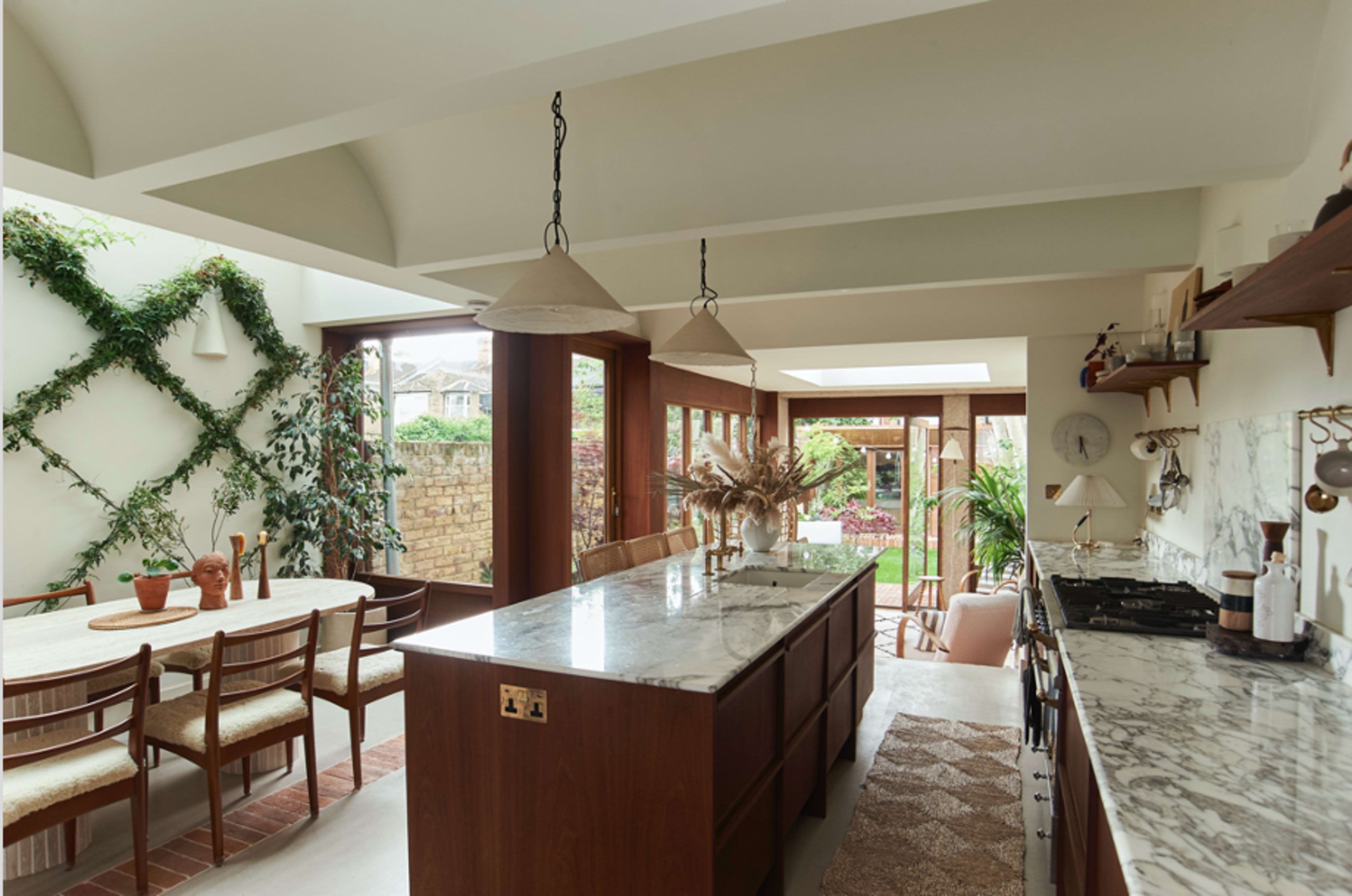 The image shows a bright and modern kitchen with an island, wooden cabinetry, a dining area, and large windows overlooking a garden.